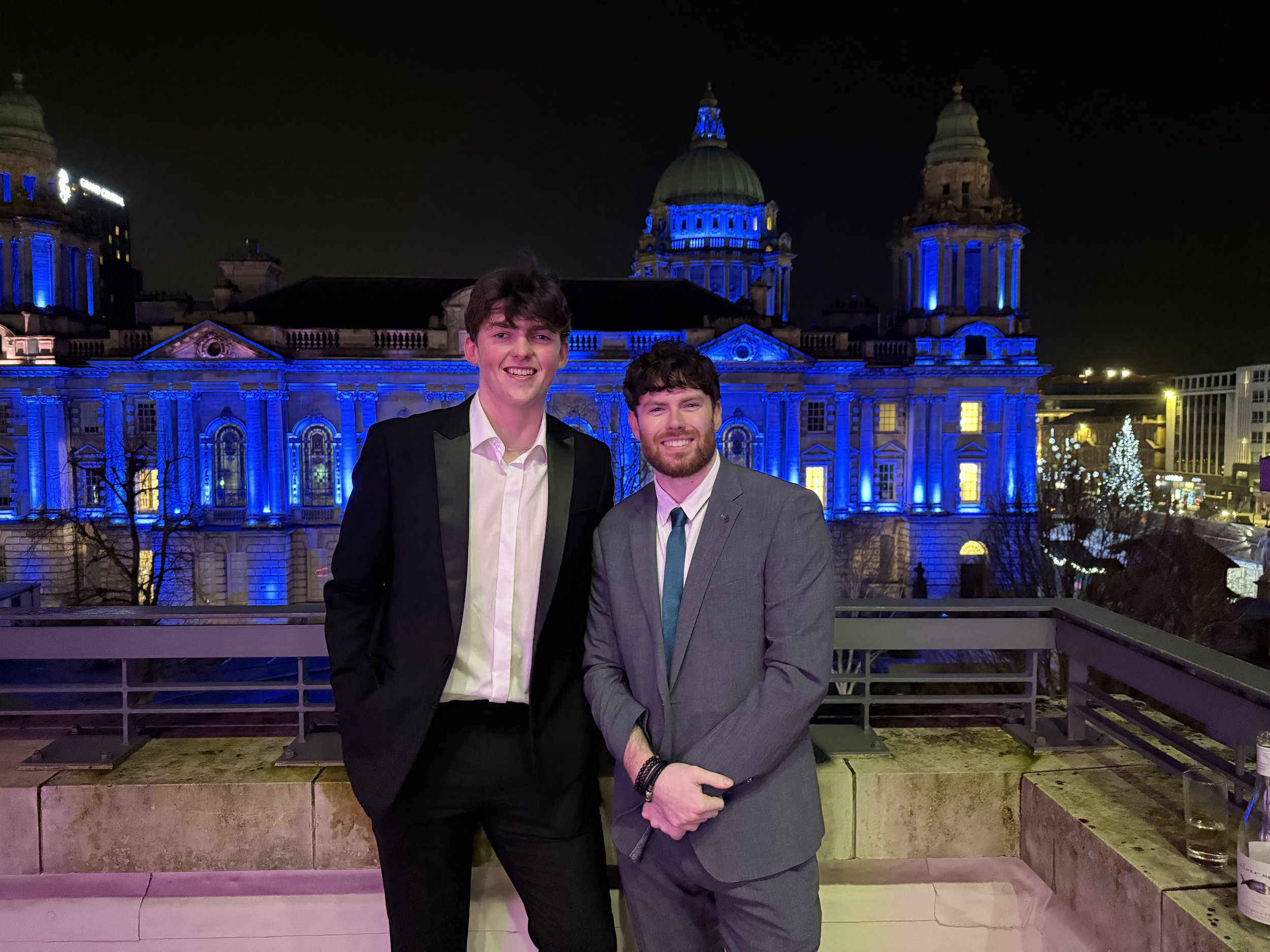 Two men in suits standing on a rooftop balcony at night, with a lit-up historic building featuring domed towers in the background.