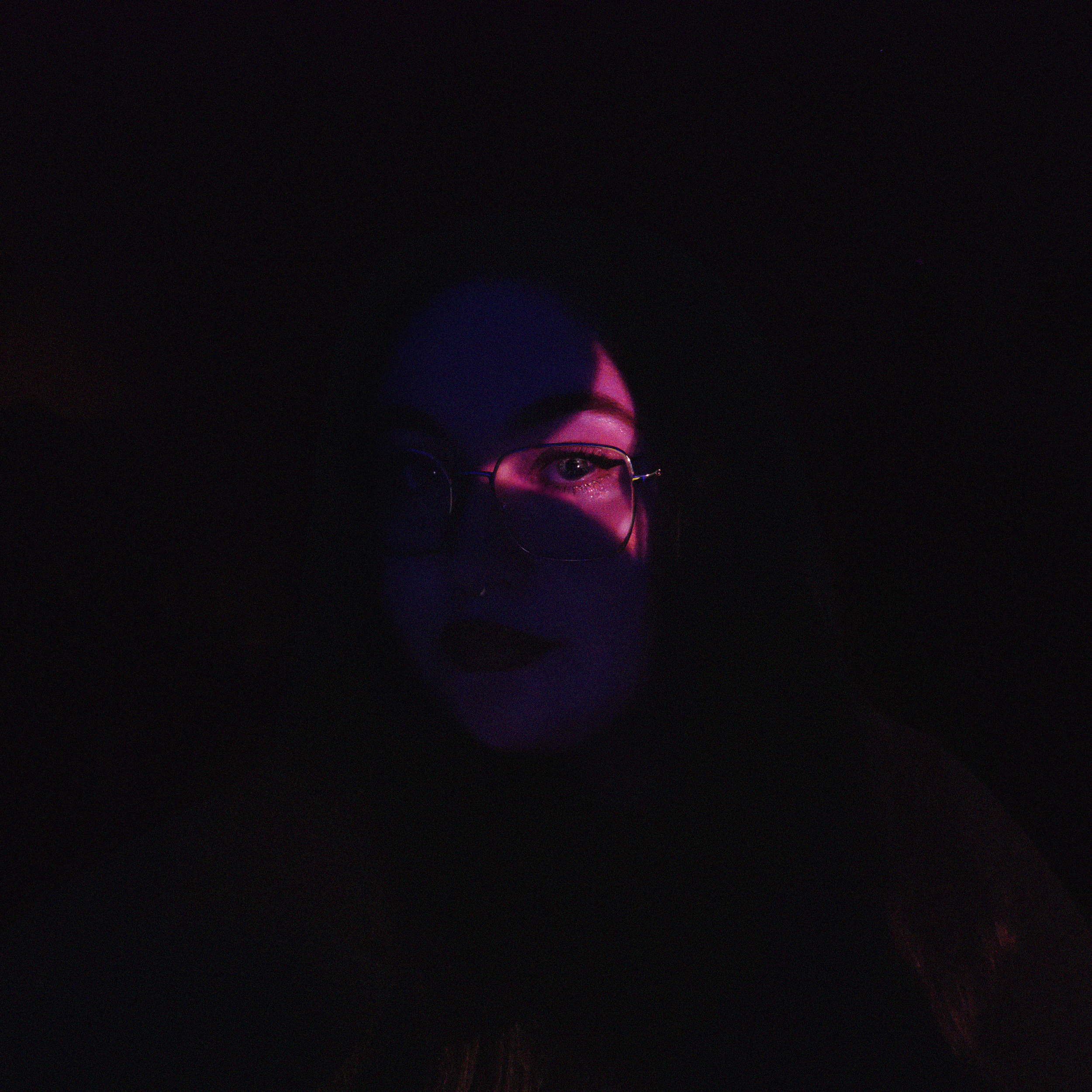 Portrait sombre d'une personne portant des lunettes, avec un éclairage violet et rose partiel sur le visage, dans un environnement obscur.