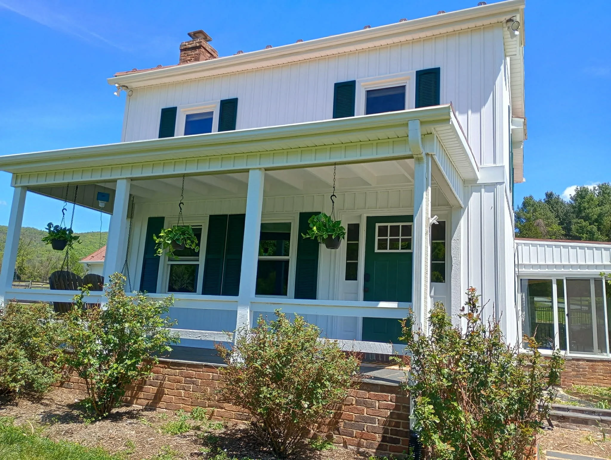 Rear view of a white two-story house with a porch, green shutters, hanging plants, and a brick foundation under a blue sky.