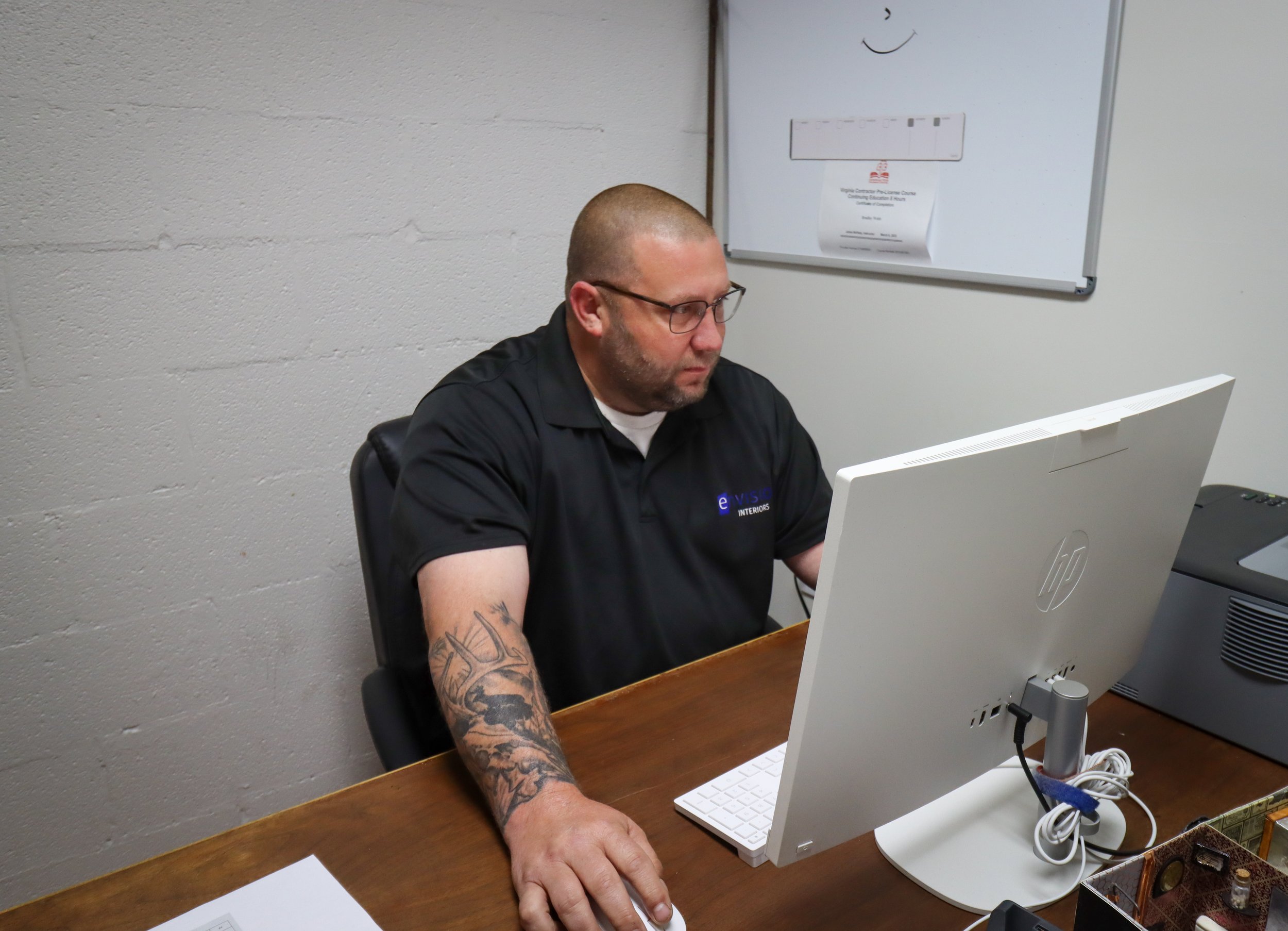A man with glasses, a beard, and tattoos on his left arm, sitting at a wooden desk and working on a white desktop computer in an office with a white brick wall and a whiteboard in the background.