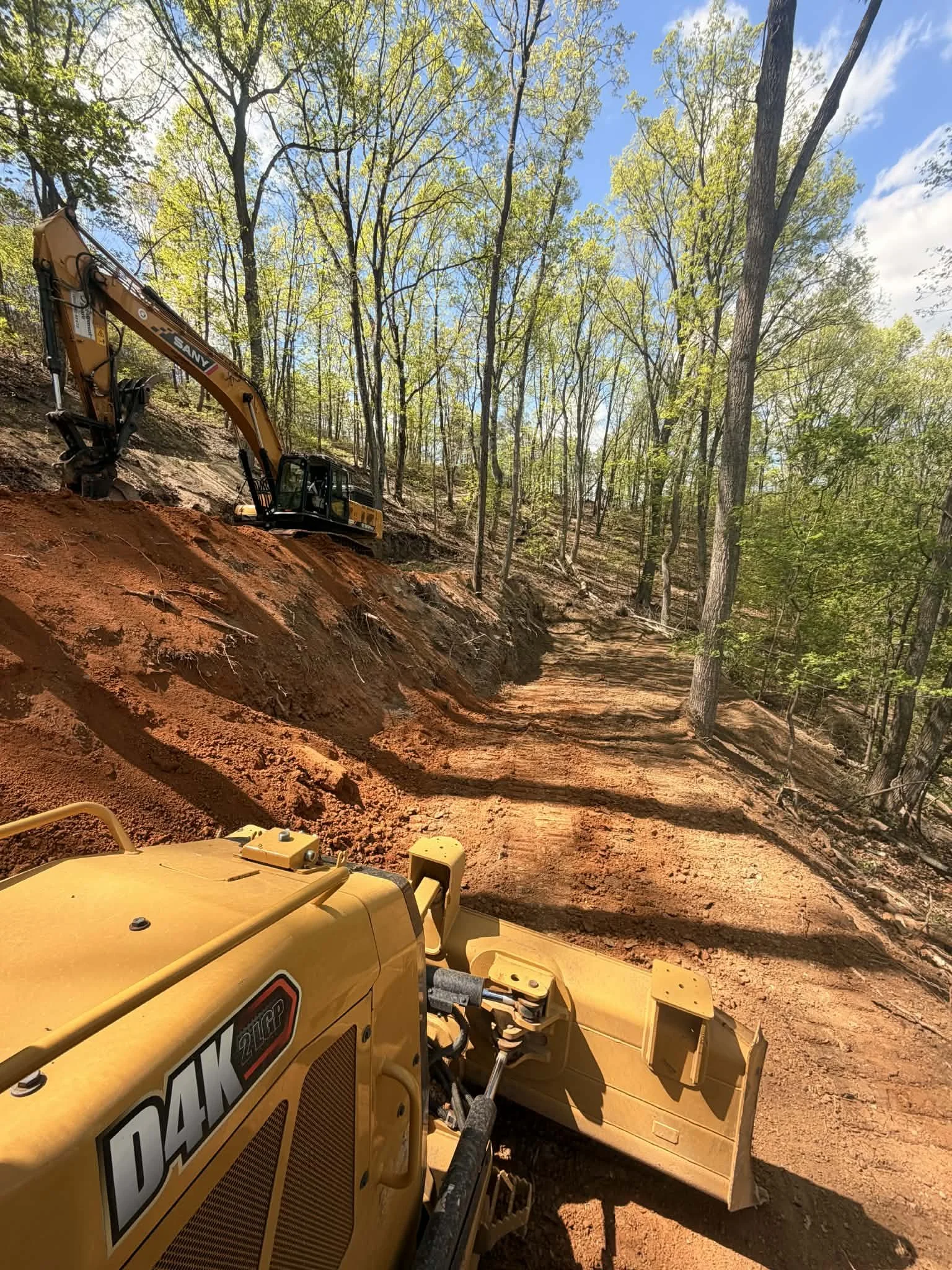 A yellow Caterpillar excavator working on a dirt trail in a wooded area with tall trees and blue sky.
