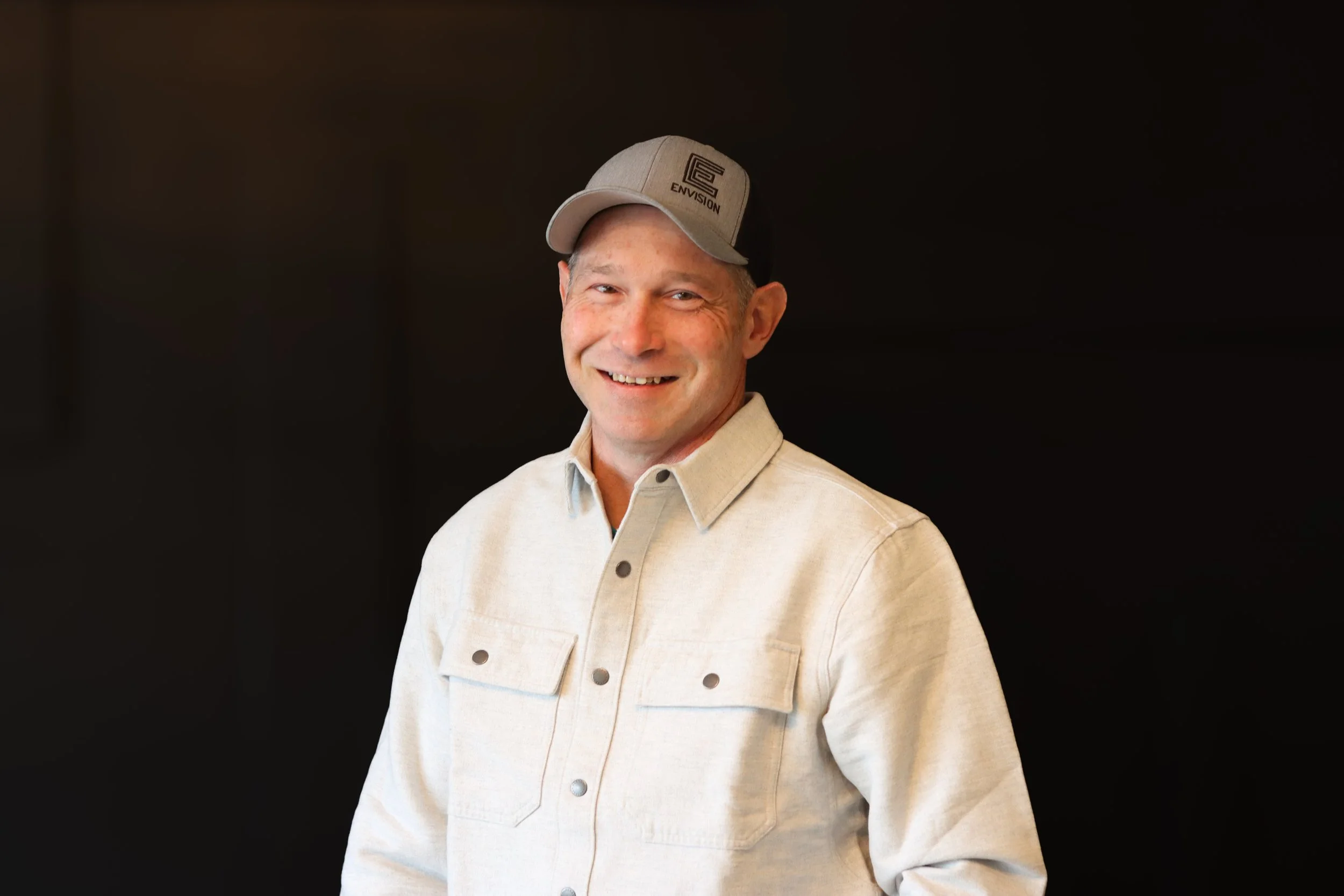 Portrait of a smiling man wearing a beige cap and a light-colored button-up shirt, standing against a dark background.