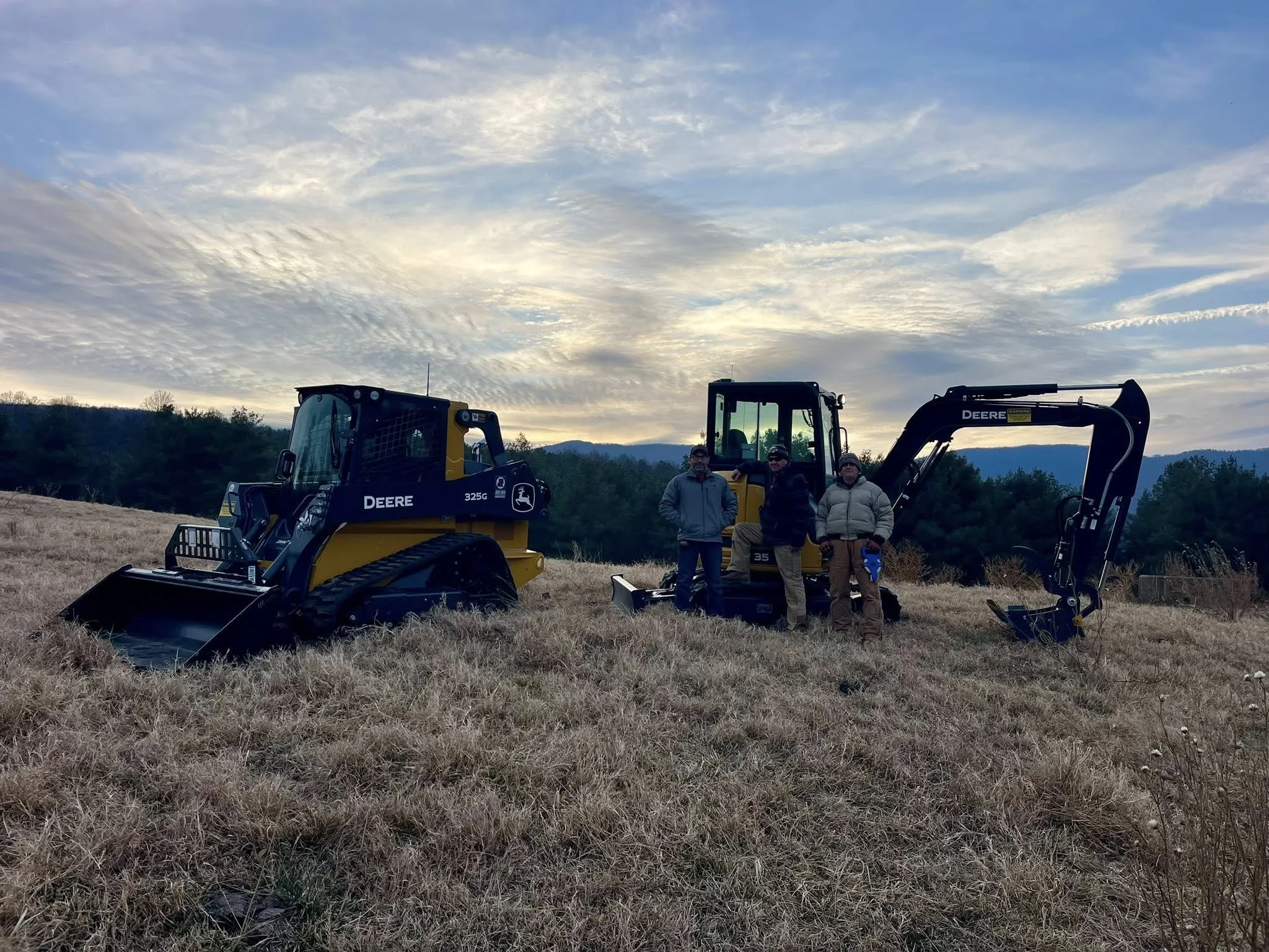 Three people are standing in a field with two pieces of yellow construction equipment, a John Deere skid steer and an excavator, under a cloudy sky with a background of trees and mountains.