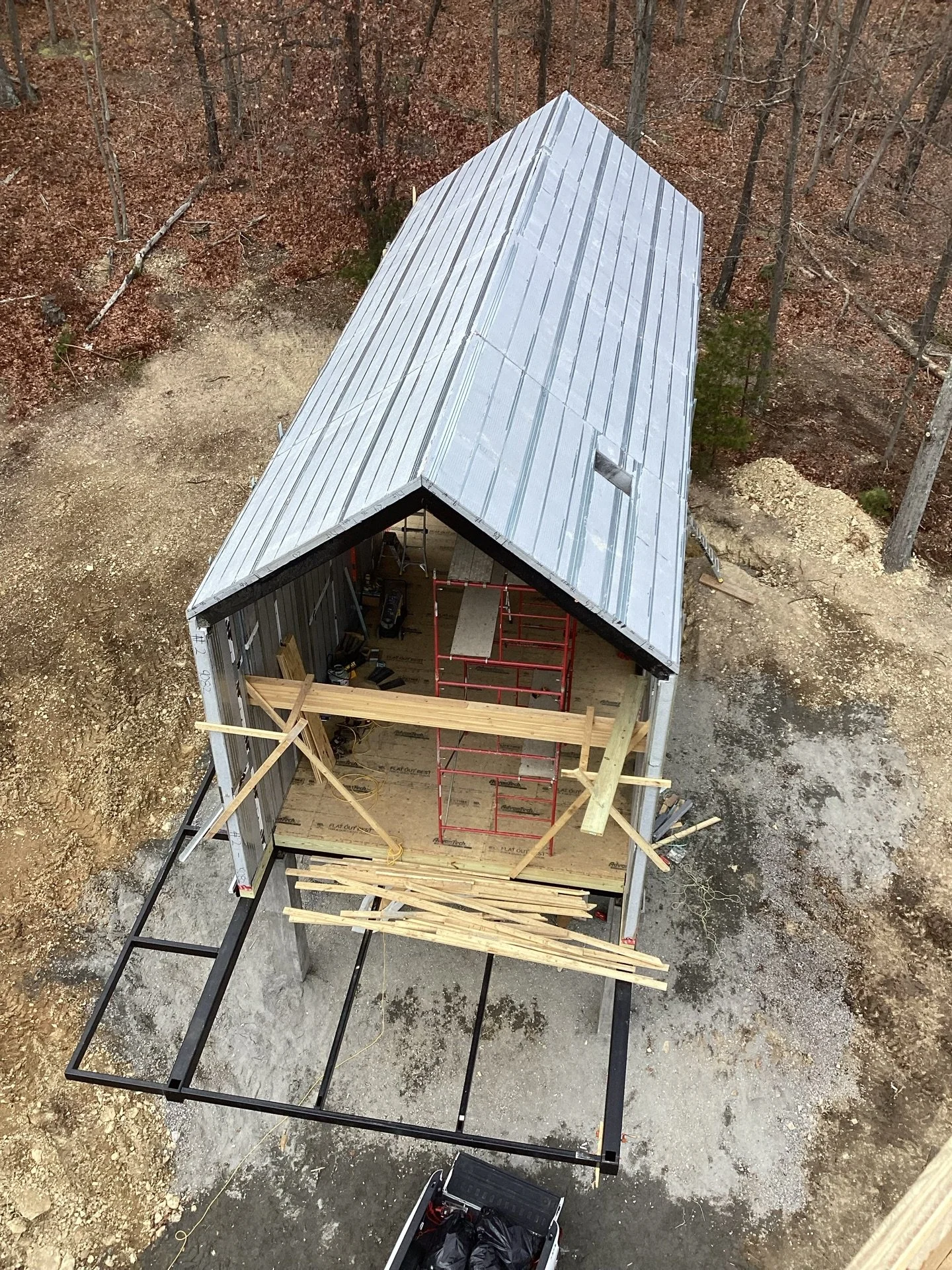 Aerial view of a construction site with a partially built small building, showing a metal roof and wooden framing, surrounded by trees and dirt ground.