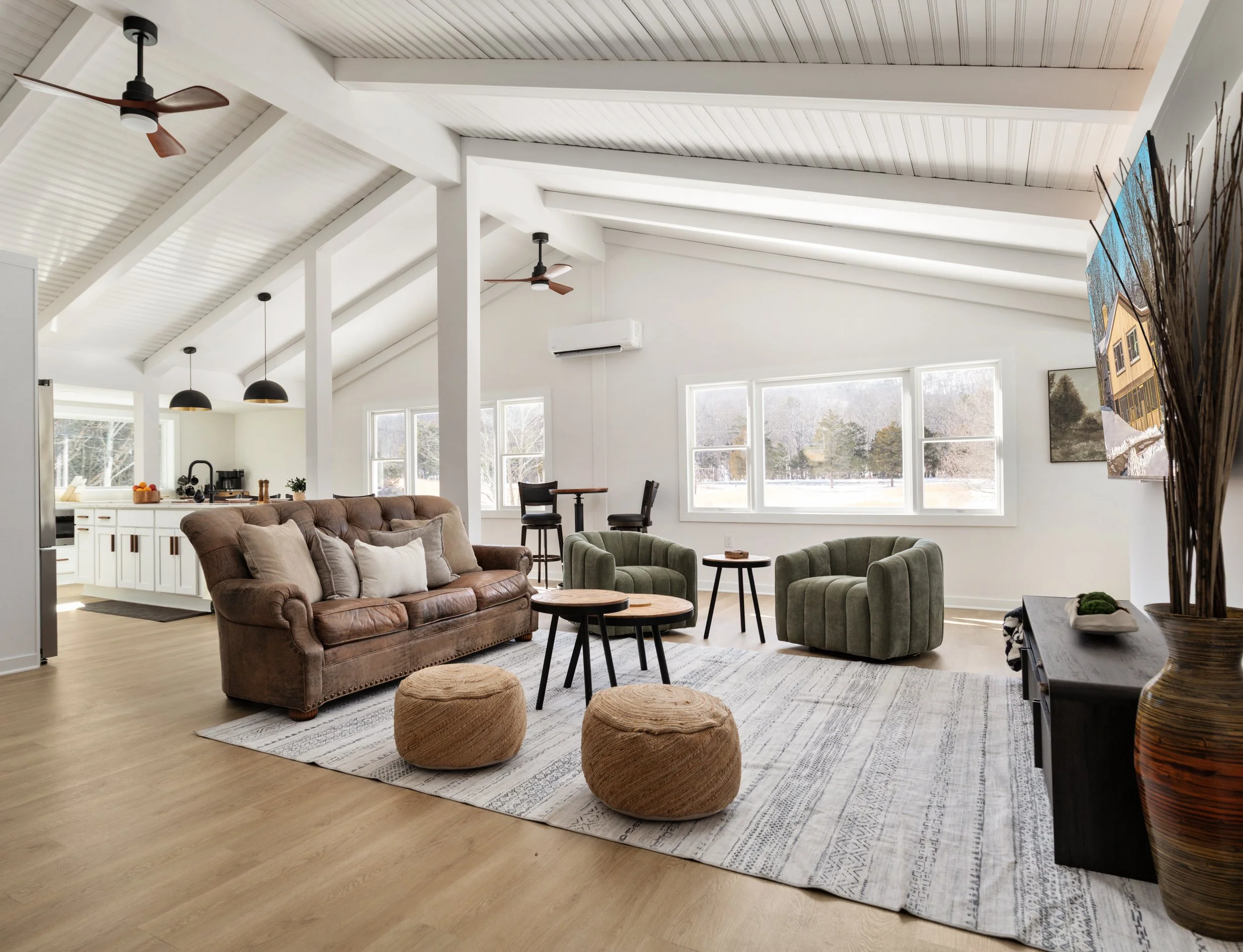 Open concept living room with high vaulted ceilings, white walls, and large windows showing trees outside. Contains a brown tufted leather sofa, green armchairs, a patterned rug, and natural fiber poufs. Kitchen area visible in the background with white cabinetry and black appliances. Ceiling fans and light fixtures hang from the ceiling.