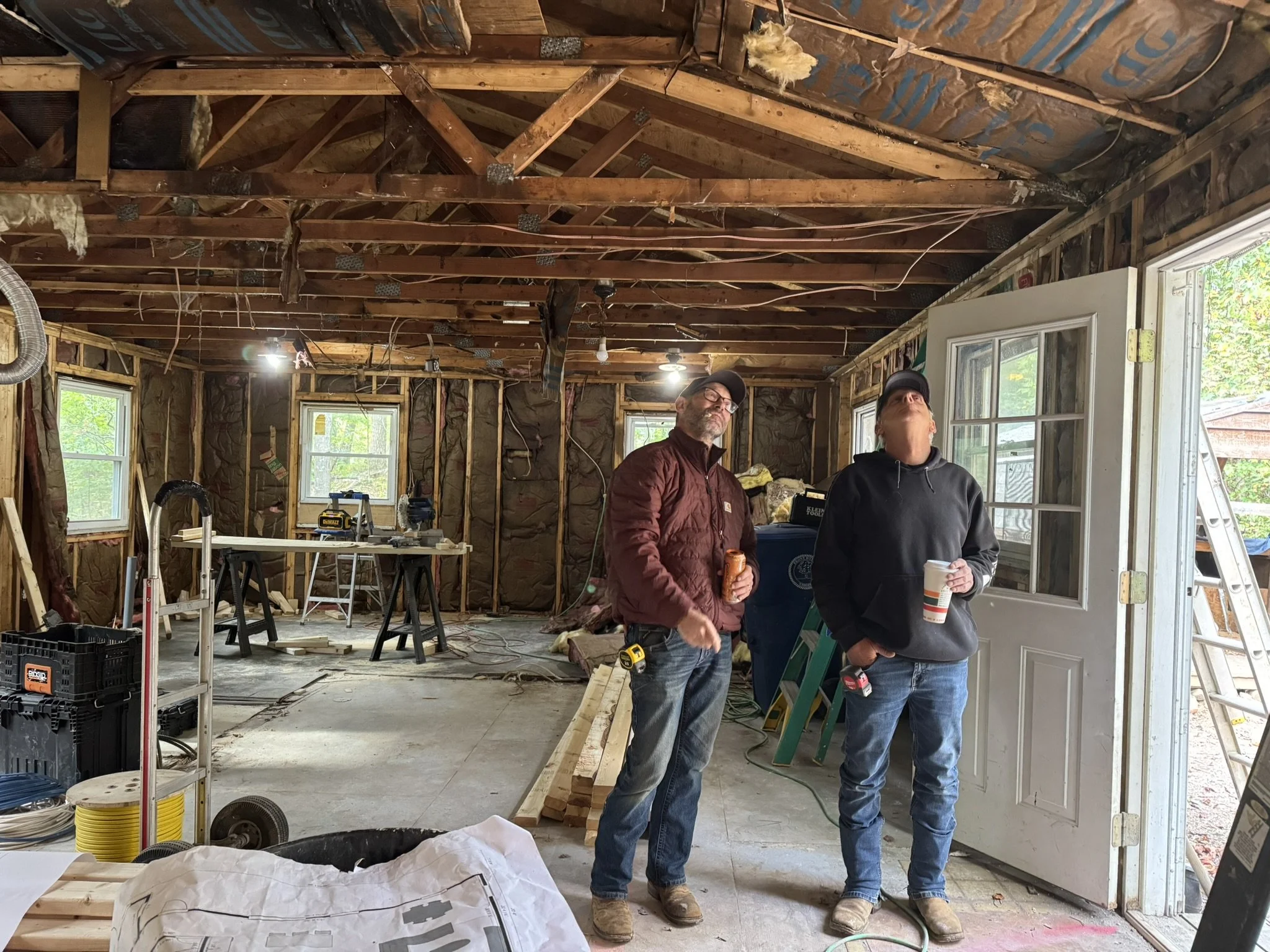 Two men stand inside a house under construction or renovation, with unfinished walls, exposed wooden beams, wiring, and construction tools around them. They are holding drinks and looking up at the ceiling.