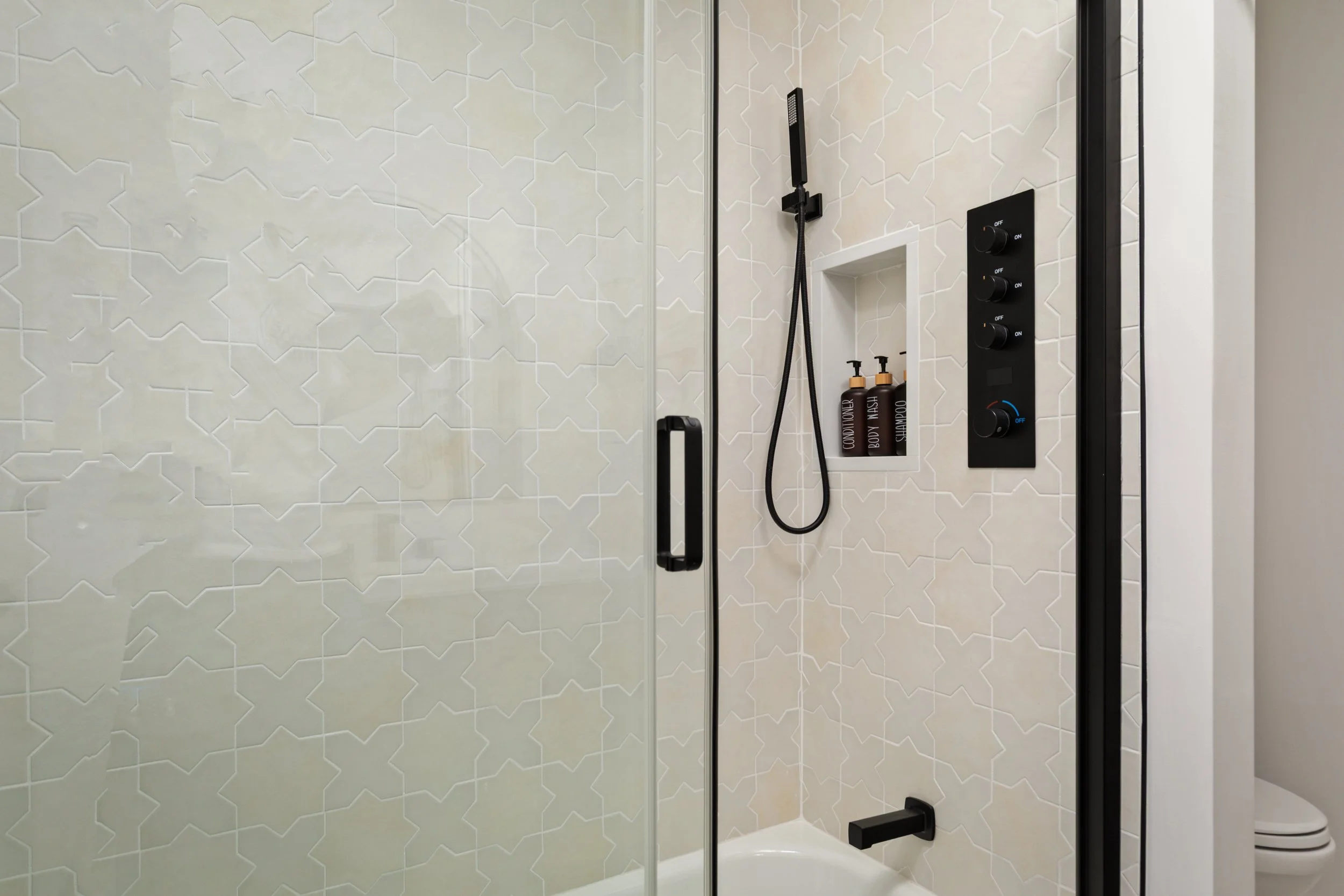Modern shower with a black frame, white geometric patterned tiles, a black handheld showerhead on a rail, a built-in shelf with shampoo bottles, and a walk-in toilet in the corner.