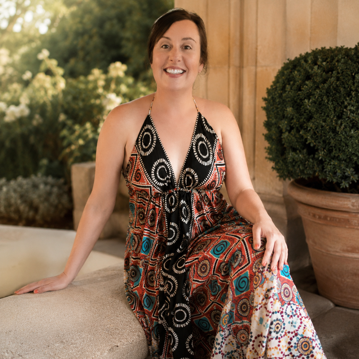 A woman with short brown hair smiling and wearing a colorful patterned halter dress sitting outdoors on a stone bench surrounded by greenery.