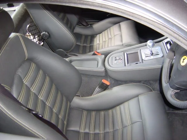 The interior of a Ferrari car showing two black and grey leather seats with yellow stitching, a centre console with gear shifter, and a passenger seat with a window net behind it.