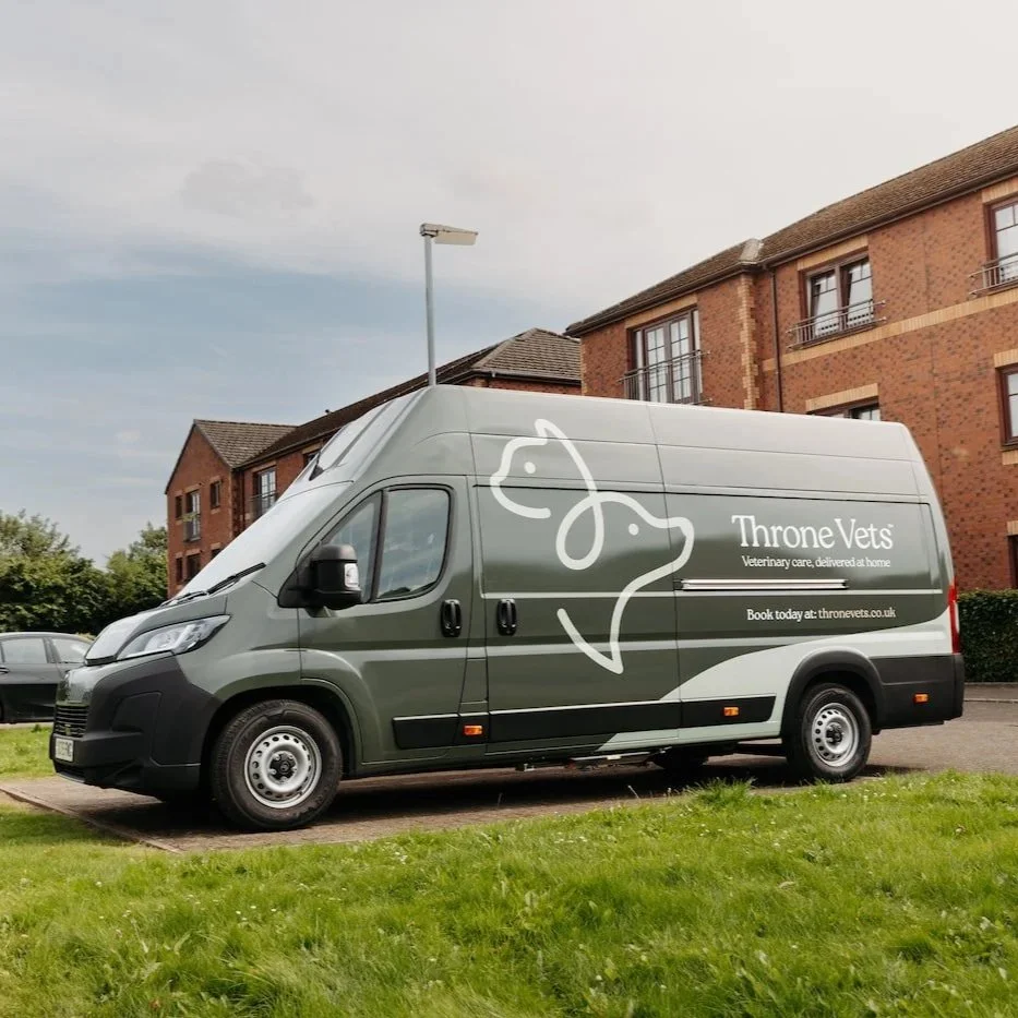 A veterinary mobile clinic vehicle for Throne Vets parked outdoors on a paved area next to green grass with multi-story brick apartment buildings in the background.