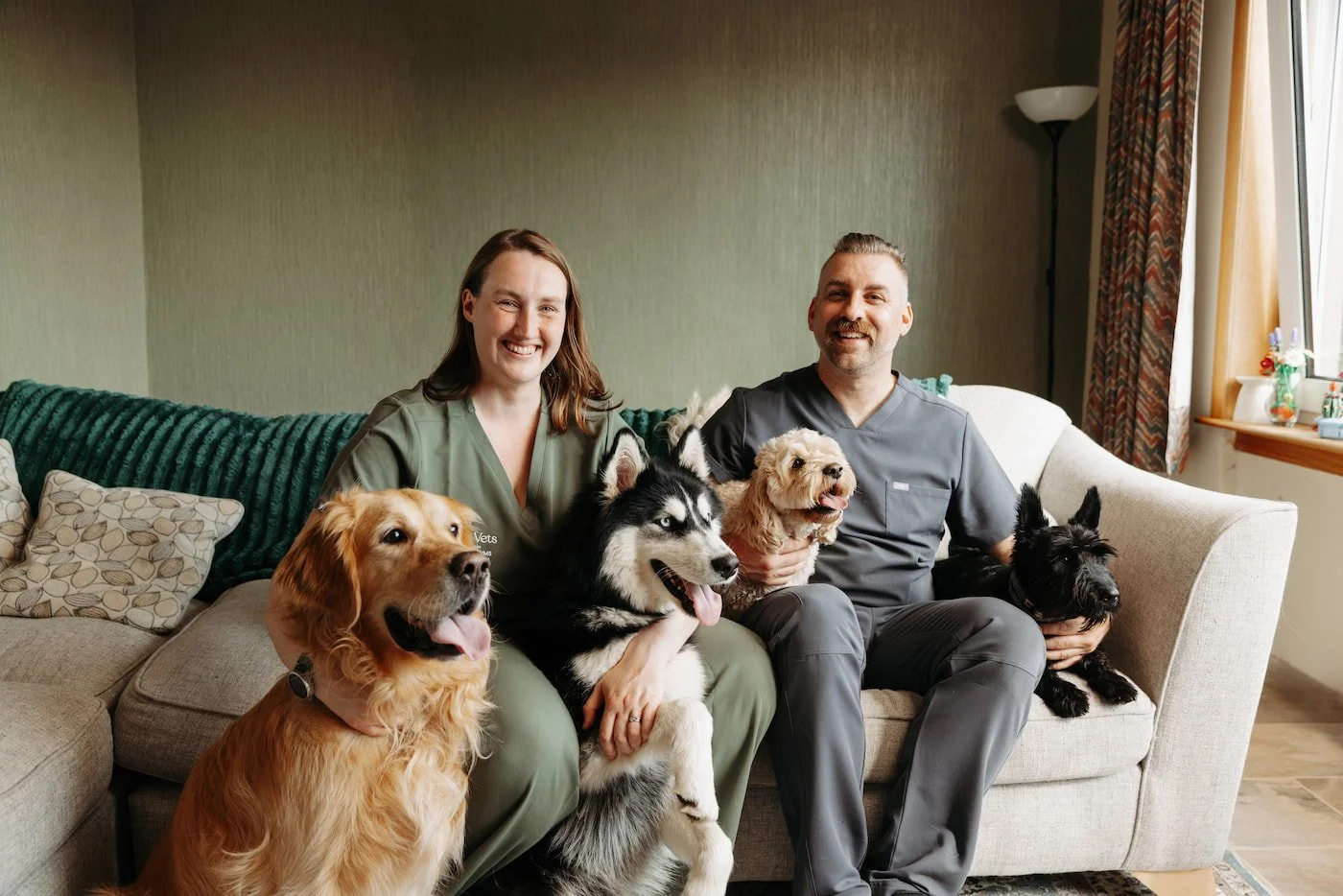 Two veterinary professionals, Rachel and David from Throne Vets, sitting on a sofa smiling with six dogs in a living room.