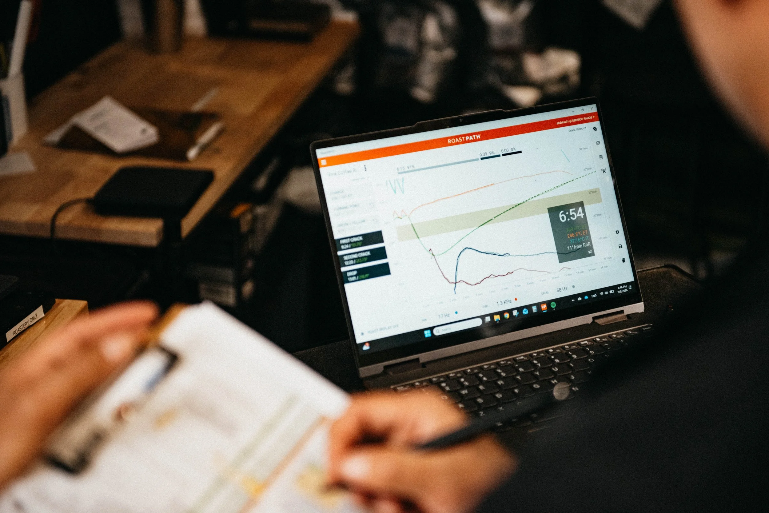 A person holding a notepad with notes, with a laptop on a wooden table displaying a graph with multiple lines and data points.