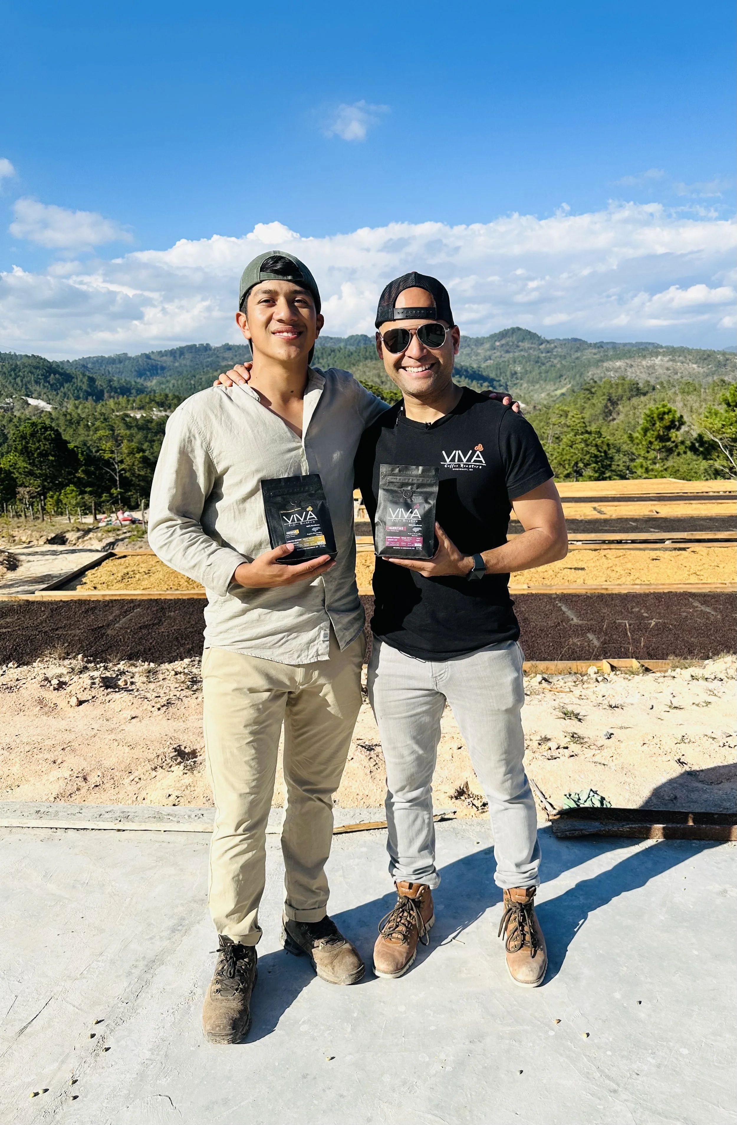 Two men standing outdoors on a sunny day, holding bags of coffee, with mountains and blue sky in the background.