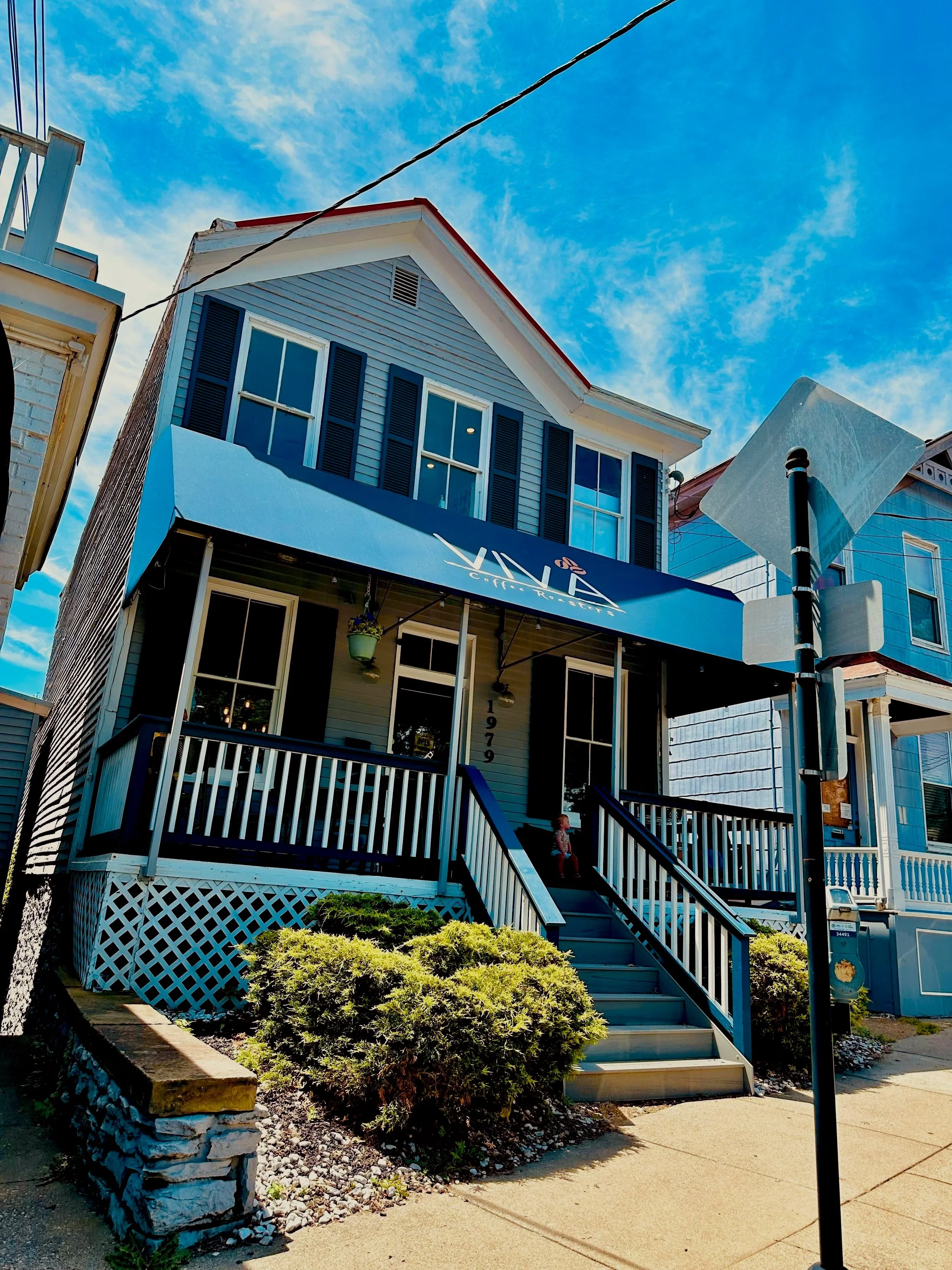 A two-story house with a blue exterior, black shutters, and a blue awning over the porch, with steps leading up to the entrance. There are bushes in front and a sidewalk, with a street sign and another house visible nearby.