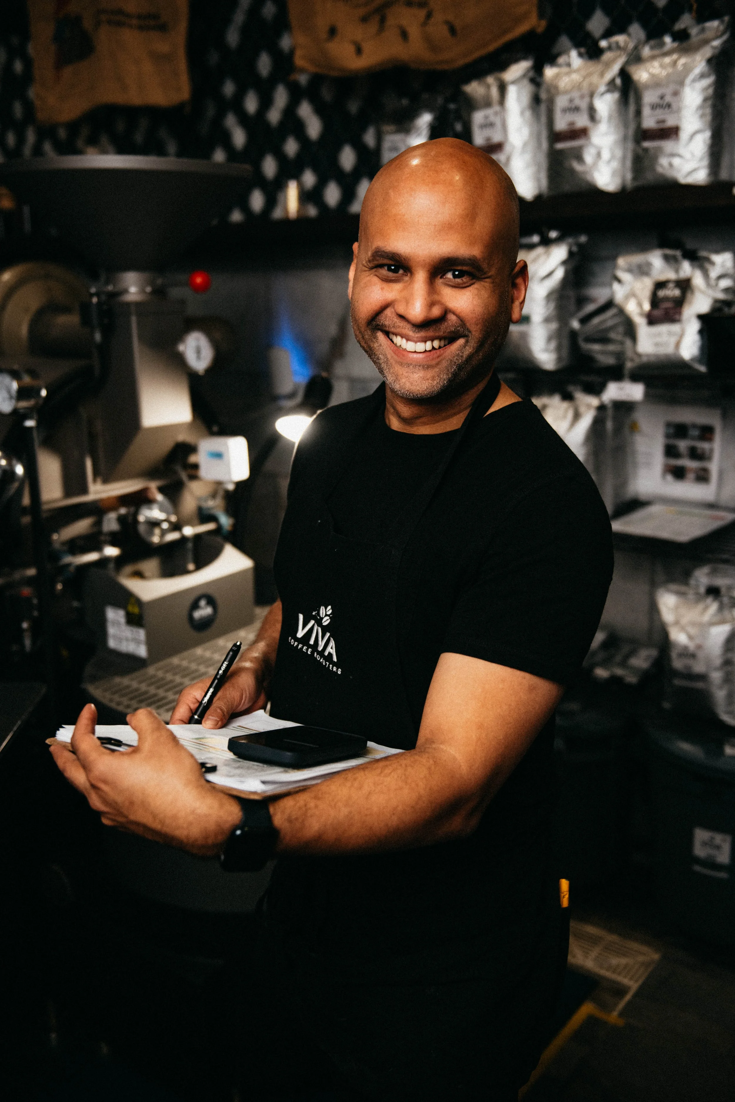 Smiling man in a black t-shirt and apron working in a coffee shop, holding a clipboard and pen, with coffee equipment and bags of coffee beans in the background.