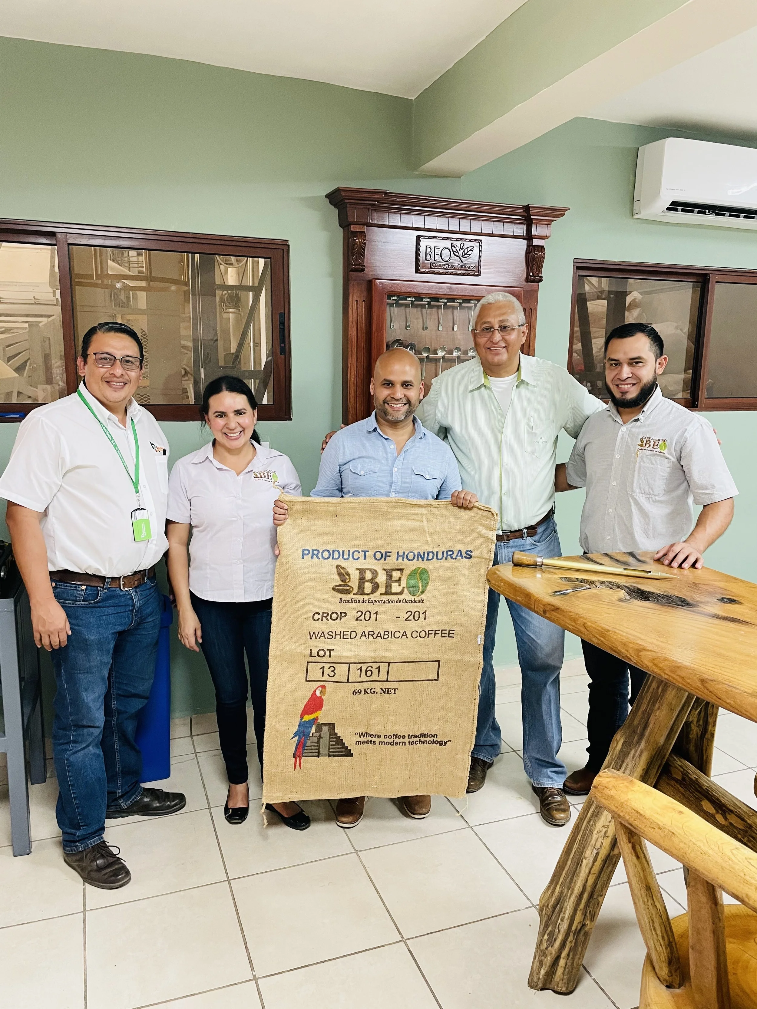 Group of five people standing indoors, holding a large coffee burlap sack with for Honduras coffee. The sack displays the words 'Product of Honduras' and 'Washed Arabica Coffee.' There is wooden furniture in the background and a window showing the kitchen or workshop area.