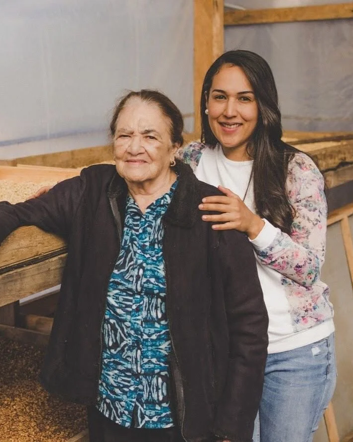 A young woman and an elderly woman standing together in a rustic setting, smiling at the camera. The young woman has long dark hair, is wearing a floral sweater and jeans, and is hugging the elderly woman from behind. The elderly woman has short hair, is wearing a patterned blouse and a dark jacket.