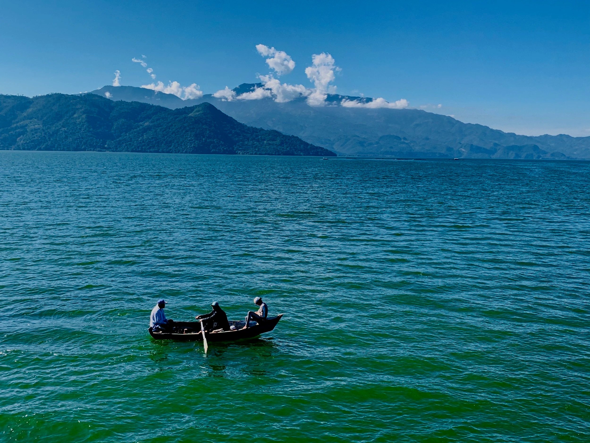 Three people in a small boat on a large lake with mountains in the background, clear blue sky, and some clouds.