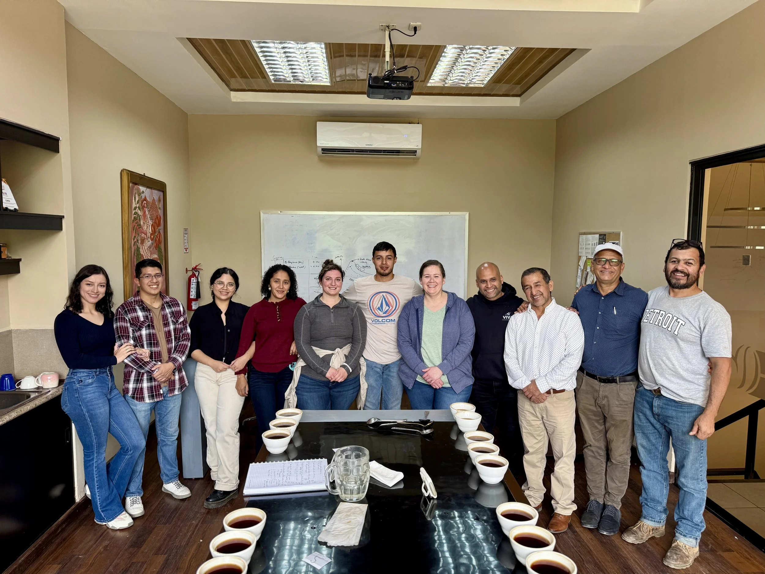 Group of twelve people standing in a classroom with a whiteboard, table with cups of coffee, and a window on the right. Some individuals are smiling and posing for the photo.