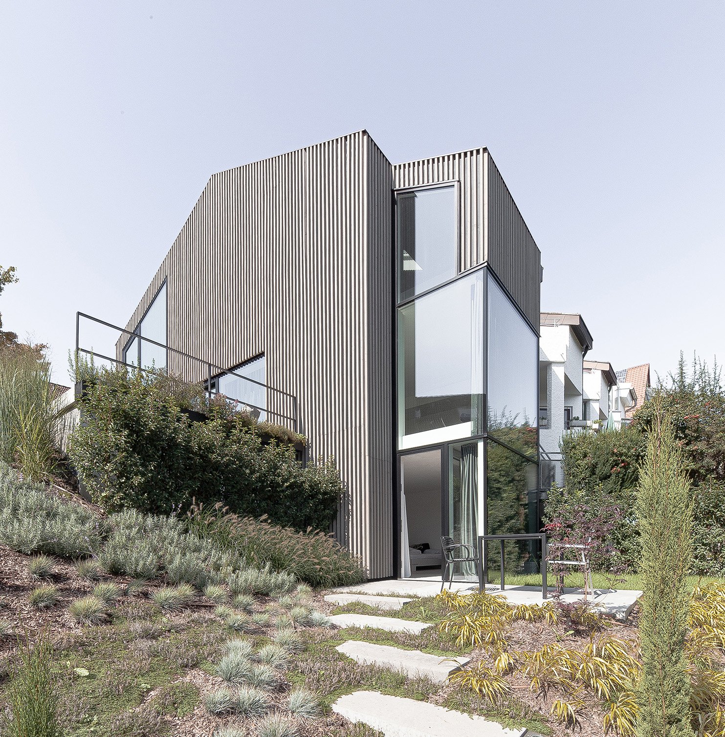 haus RU - Modernes Haus mit vertikal ausgerichtetem Metallfachwerk, große Fenster und umgebender Garten mit Steinen, Büschen und kleinen Bäumen.