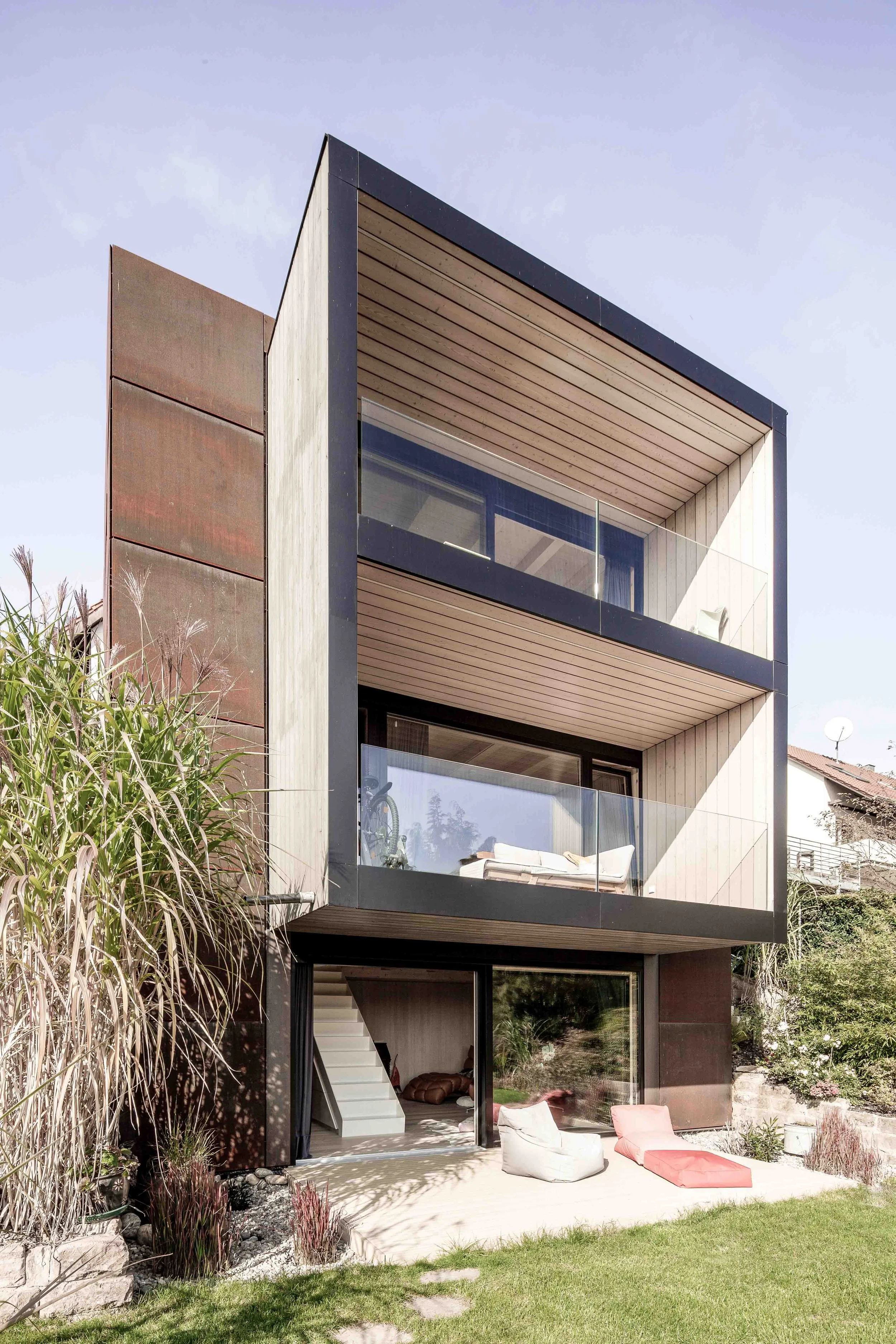 haus GRO - Moderne mehrstöckige Wohnung mit Balkon im oberen Stock, Balkon mit Glasgeländer, moderne Fassade mit Holz und Metall, Gartenterrasse mit Sitzgelegenheiten, Sonnenschirm, Pflanzen und Garten, klarer Himmel