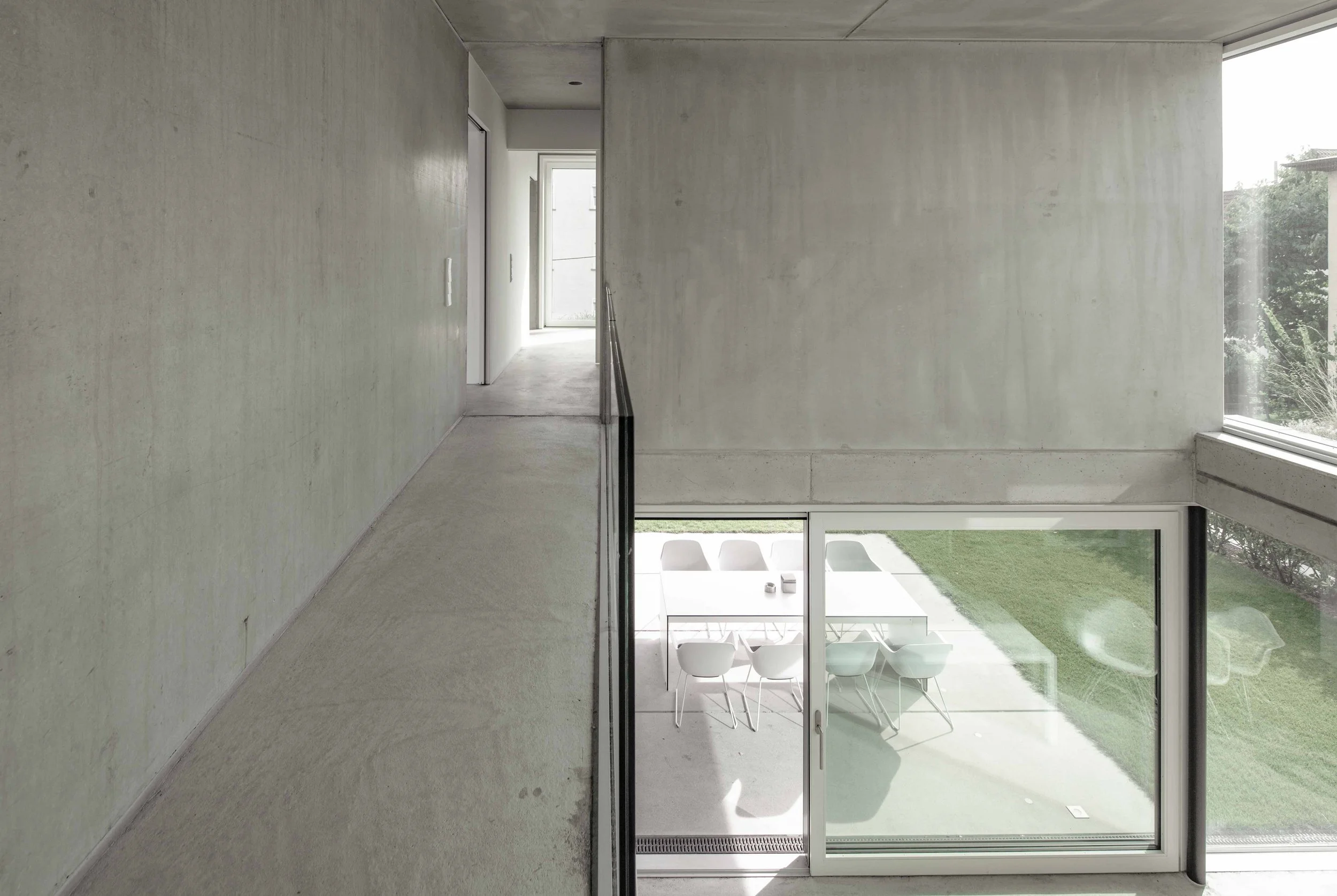 haus vO - Innenansicht eines modernen, minimalistisch gestalteten Hauses mit großen Fenstern, offener Gestaltung, sichtbaren weißen Stühlen im unteren Bereich und einem Balkon mit Blick ins Grüne.