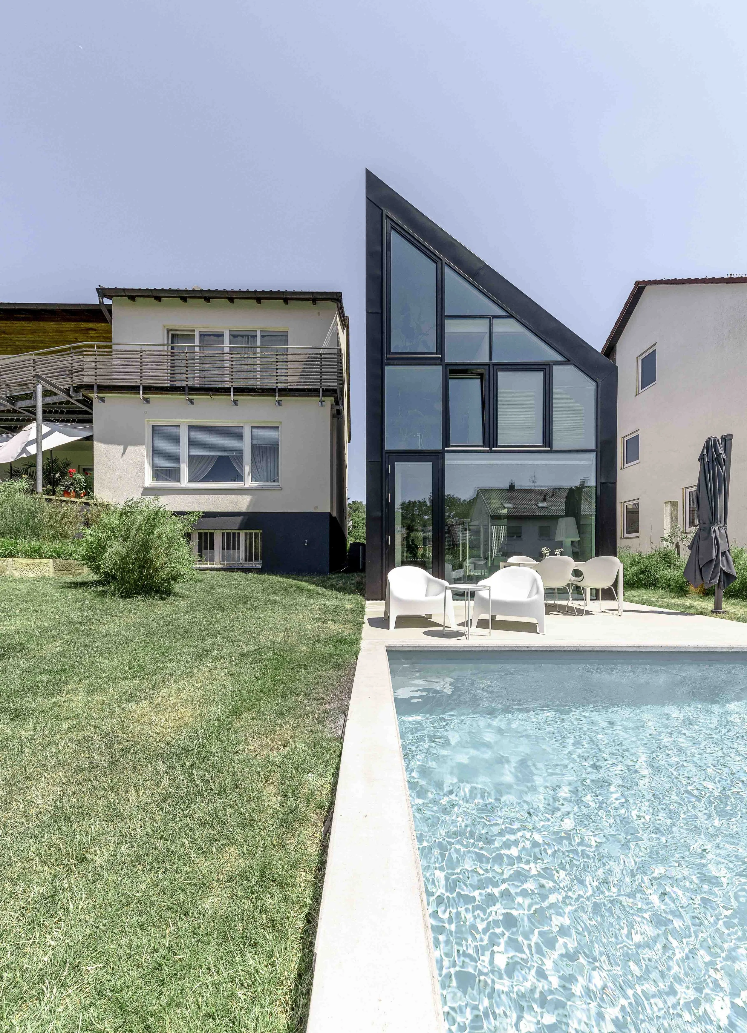 Garten mit Swimmingpool und modernen Häusern, eine Terrasse mit weißen Gartenmöbeln, ein Regenschirm und ein geschlossenes Sonnenschirm, im Hintergrund Häuser mit unterschiedlichen Fassaden, der Himmel ist bewölkt.