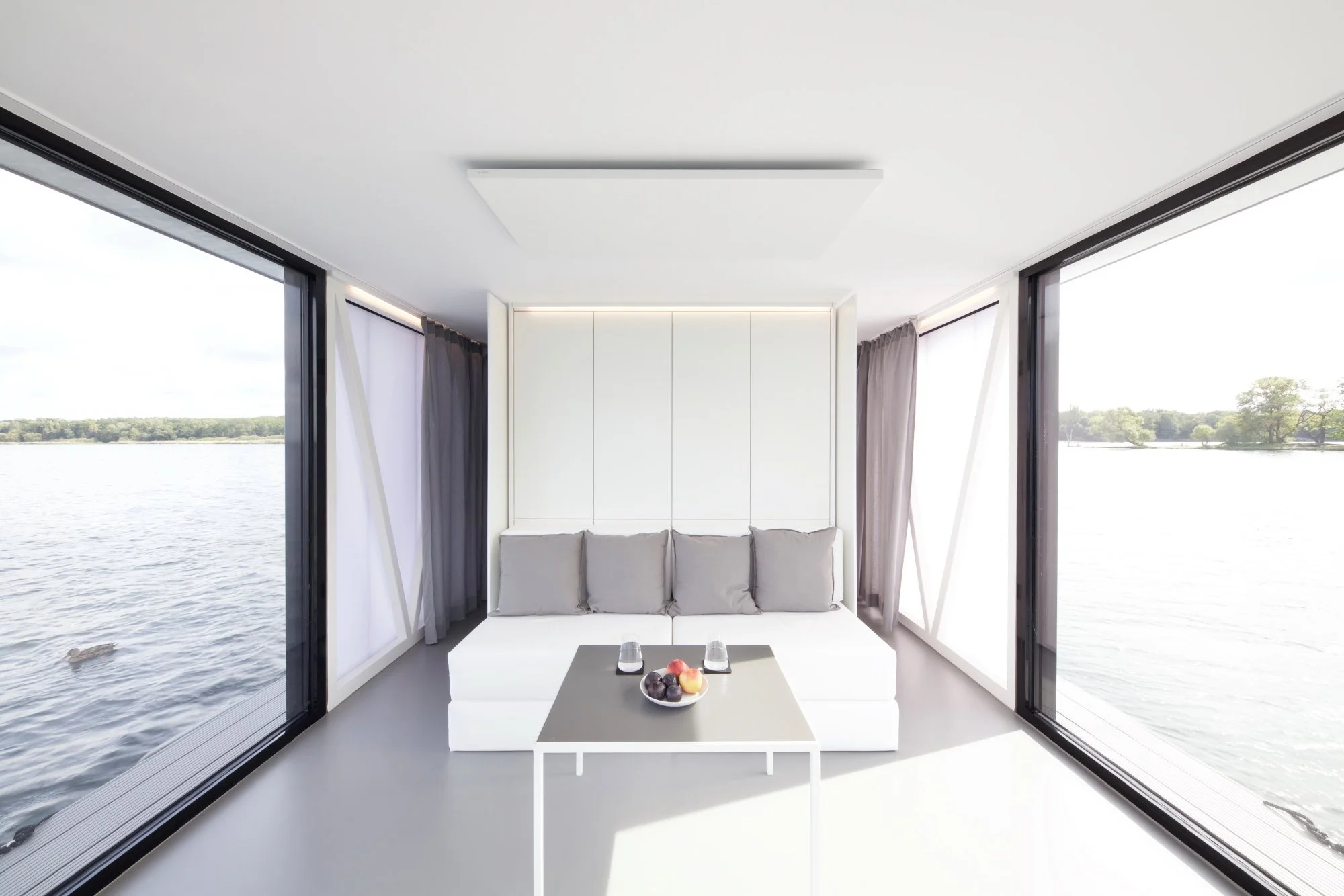 Innenraum eines modernen Raums mit großen Fenstern, Blick auf einen Fluss und Bäume, weiße Wände, graue Vorhänge, ein weißer Sitzbereich, eine kleine weiße Kaffeetafel mit Obst und Wasser.