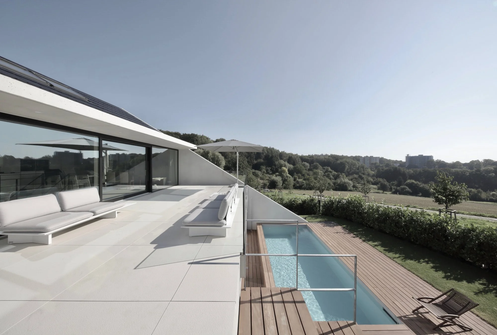haus CC - Moderne Terrasse mit weißen Sitzmöbeln, einem kleinen Pool und Blick ins Grüne bei sonnigem Himmel.