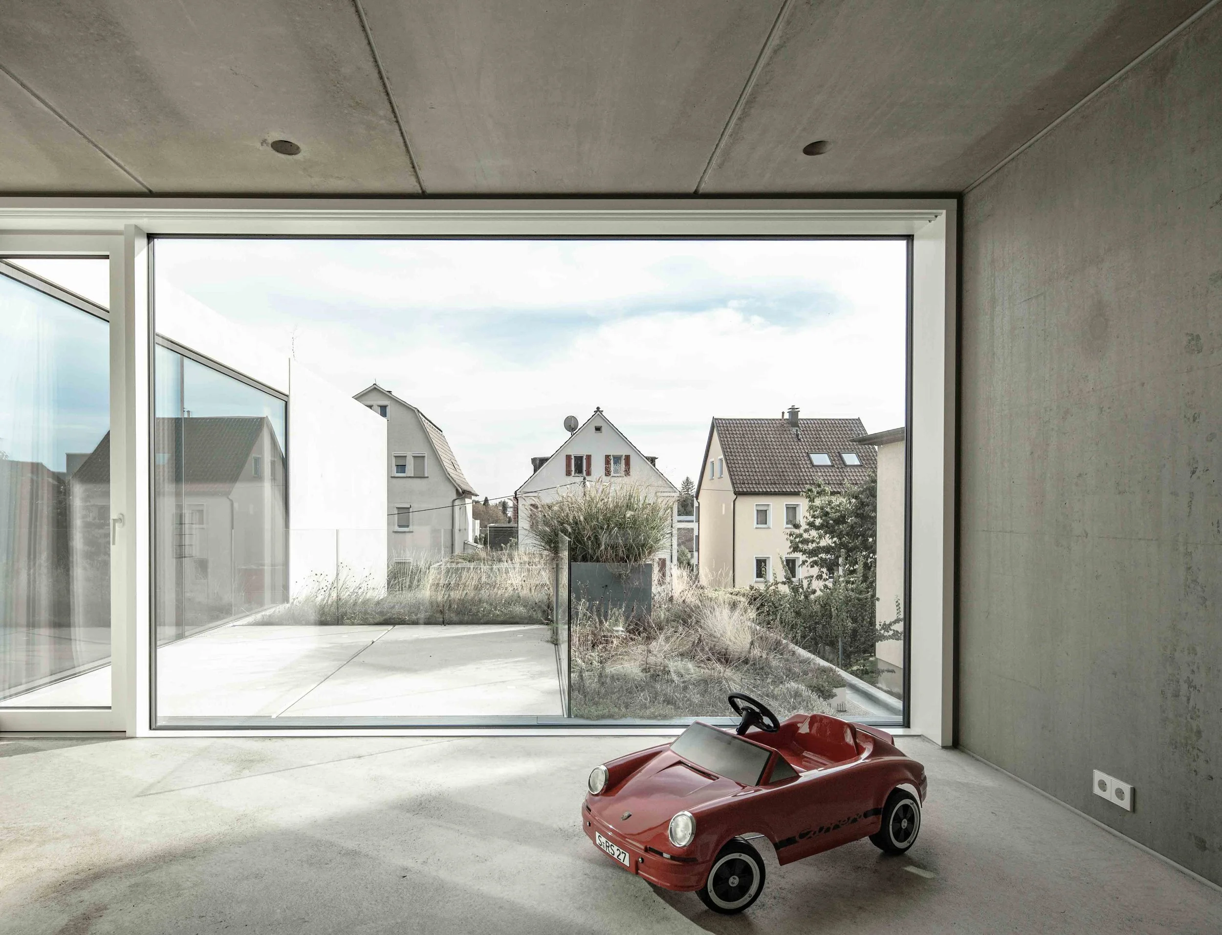 haus vO - Innenansicht eines Raumes mit großen Fenster und Blick auf Nachbarhäuser im Hintergrund, im Raum steht ein rotes Spielzeugauto.
