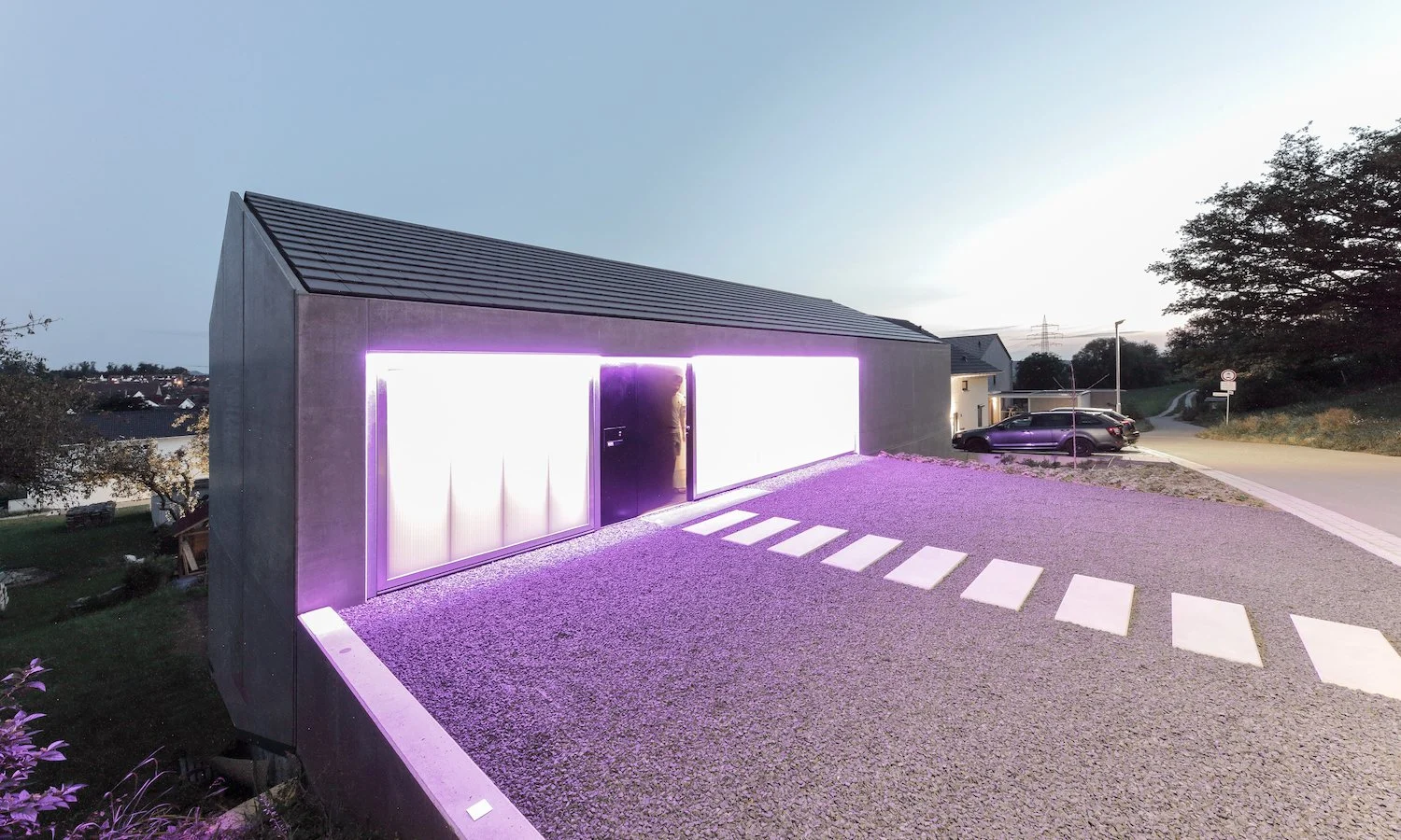 haus WÄ - Modernes Haus mit LED-Beleuchtung vor Sonnenuntergang, Parkplatz mit Autos, ländliche Umgebung mit Bäumen und Straßen.