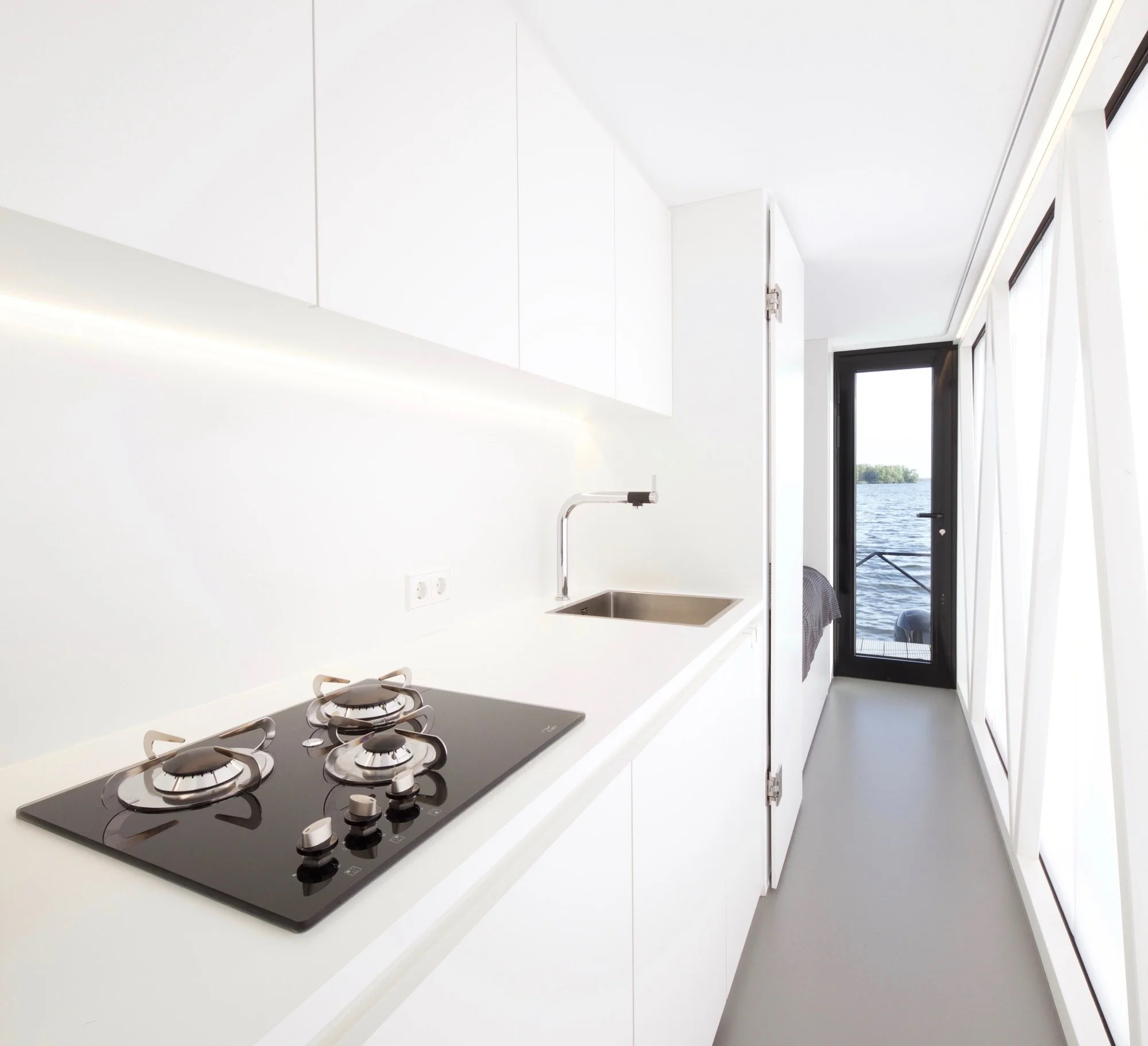 Moderne weiße Küche mit eingebautem Gasherd und Blick auf ein Fenster mit Wasseransicht im Hintergrund.