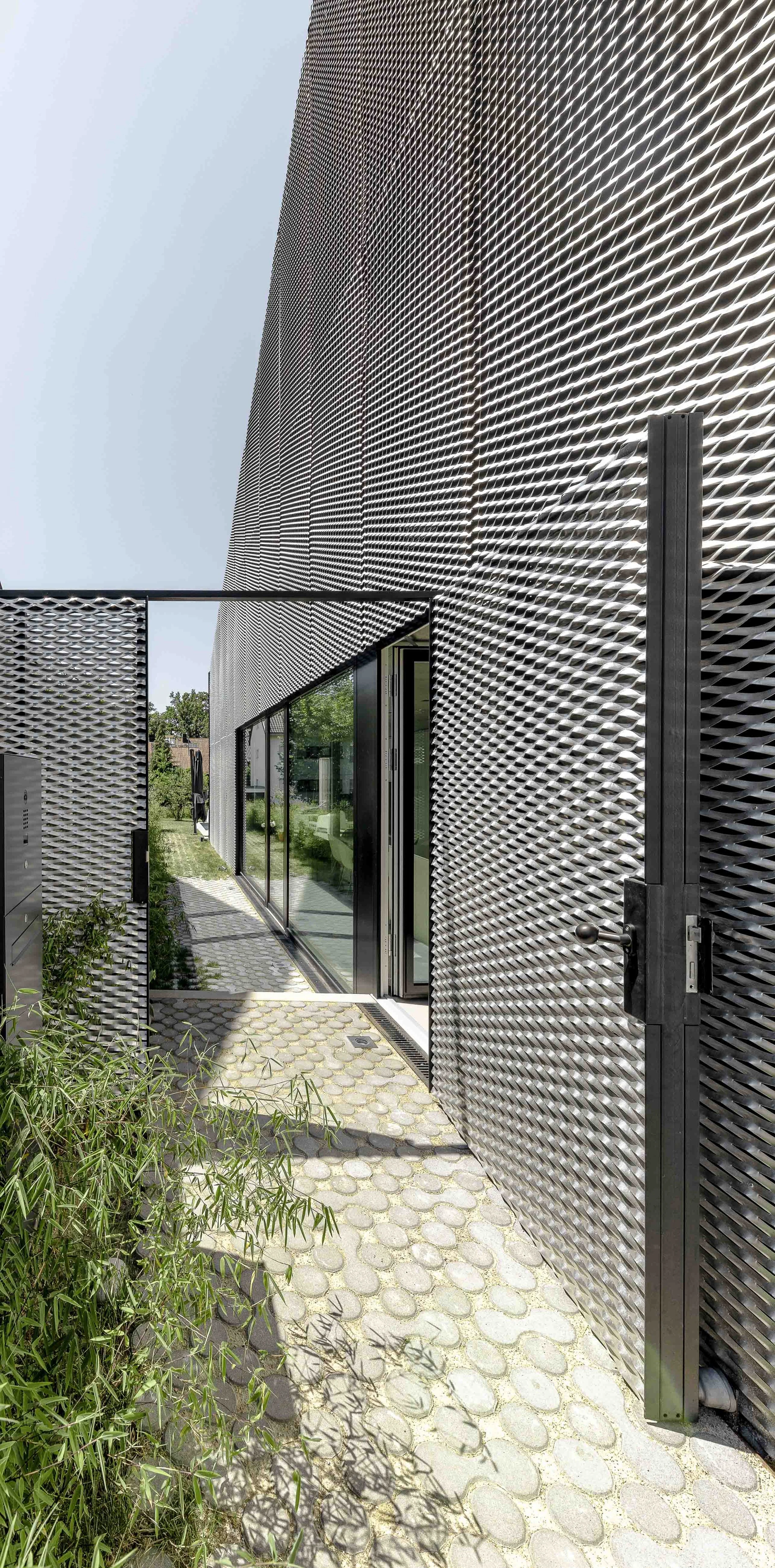 Ein modernes Gebäude mit perforierten Metallfassaden, große Glastüren und einen gepflasterten Gehweg, umgeben von Grünflächen.