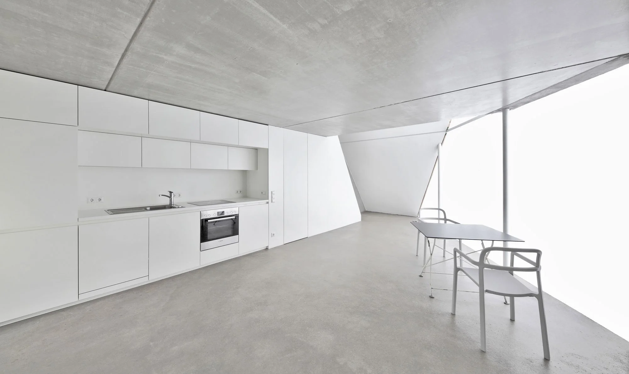 haus SO - Moderne, minimalistische Küche mit weißen Wänden, Küchenmöbeln und einem Wasserkocher, Tisch und Stühlen in einem hellen Raum mit großer Fensterfläche.