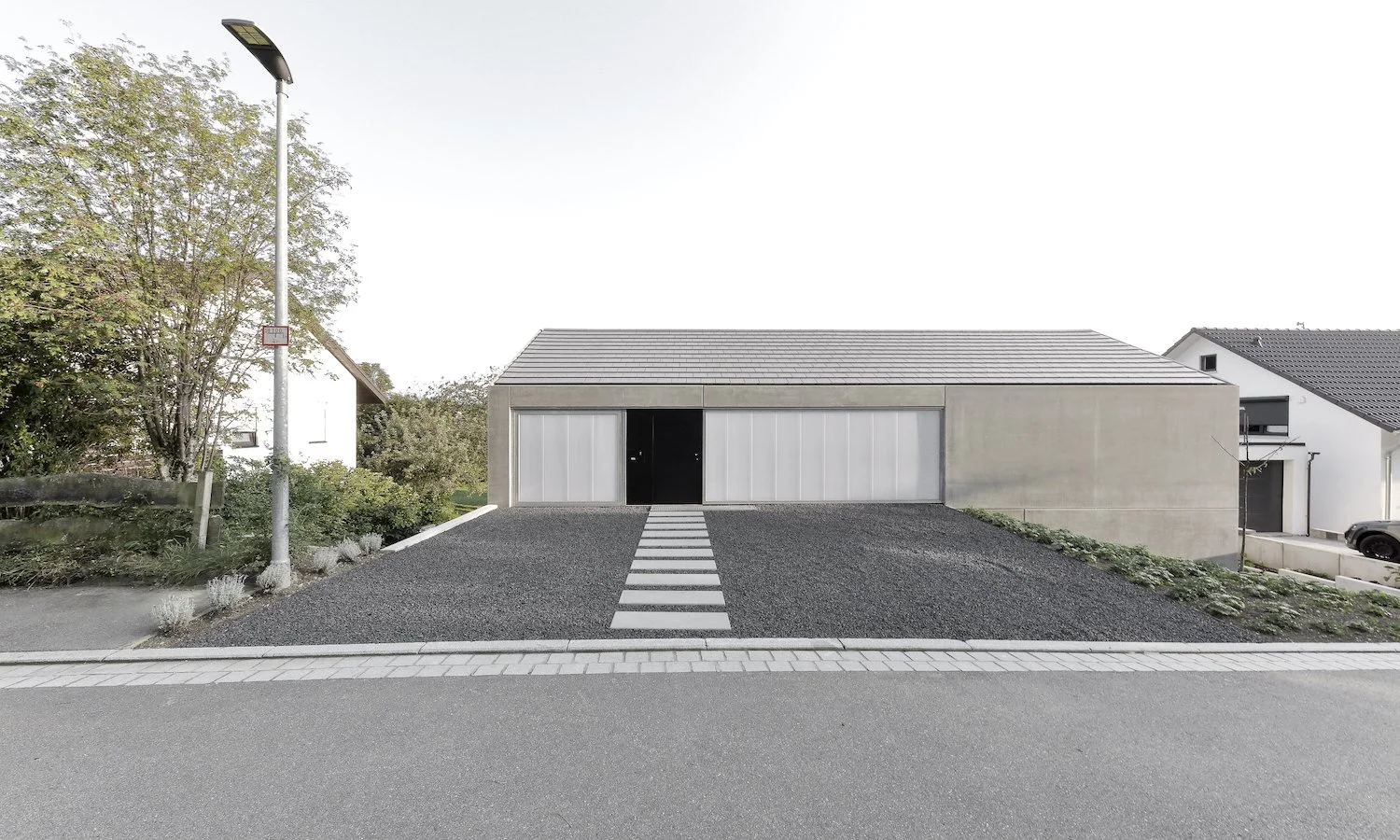 haus WÄ - Modernes, minimalistisches Haus mit grauer Fassade und Flachdach, Zebrastreifen auf der Einfahrt, umliegende Bäume und Nachbarhäuser.