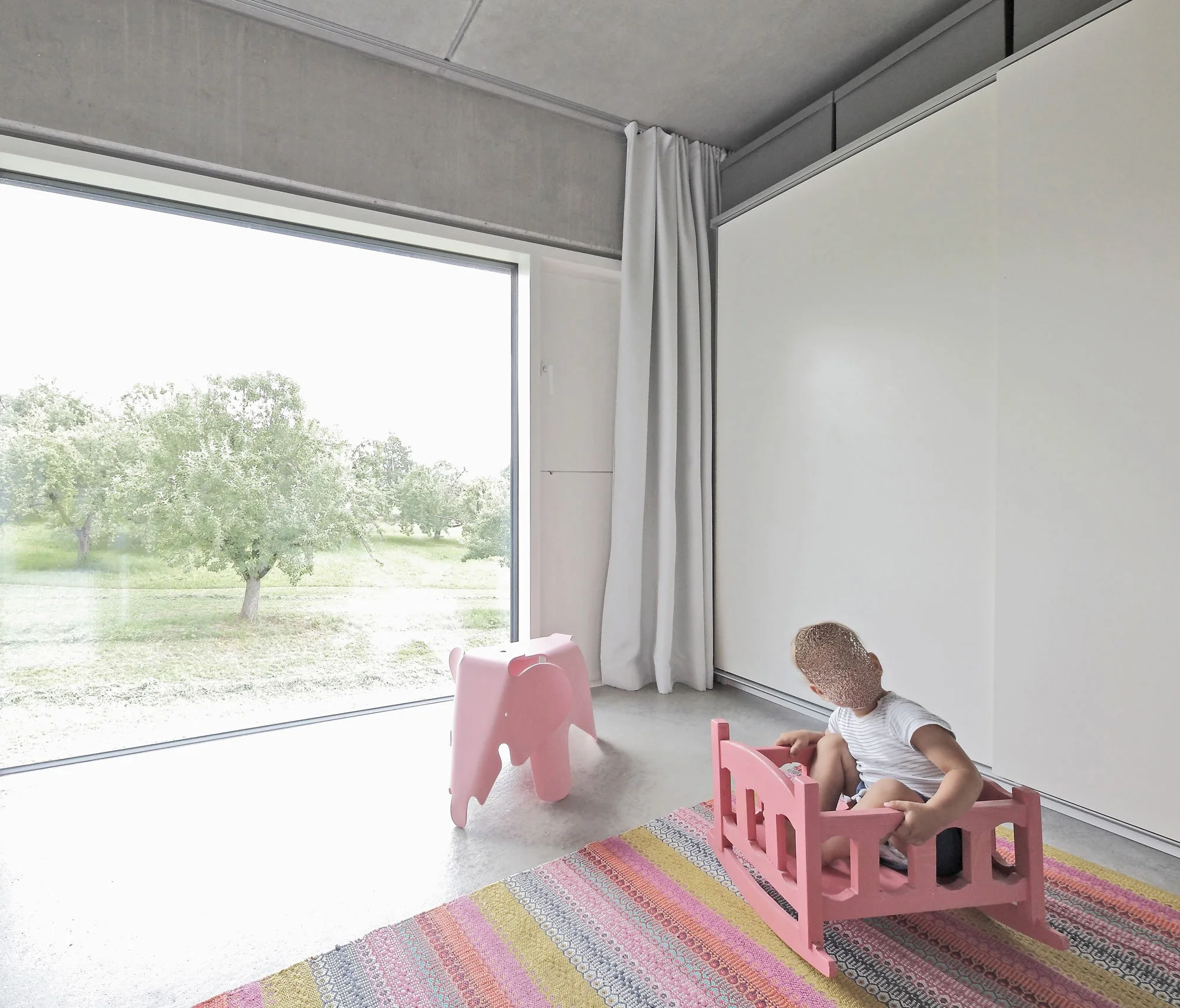 haus SG - Ein Kind spielt auf einem bunten Teppich im Innenraum mit einem pinken Spielzeugtraktor und einer pinken Elefantenfigur, vor einer großen Fensterfront mit Blick auf grüne Bäume im Freien.