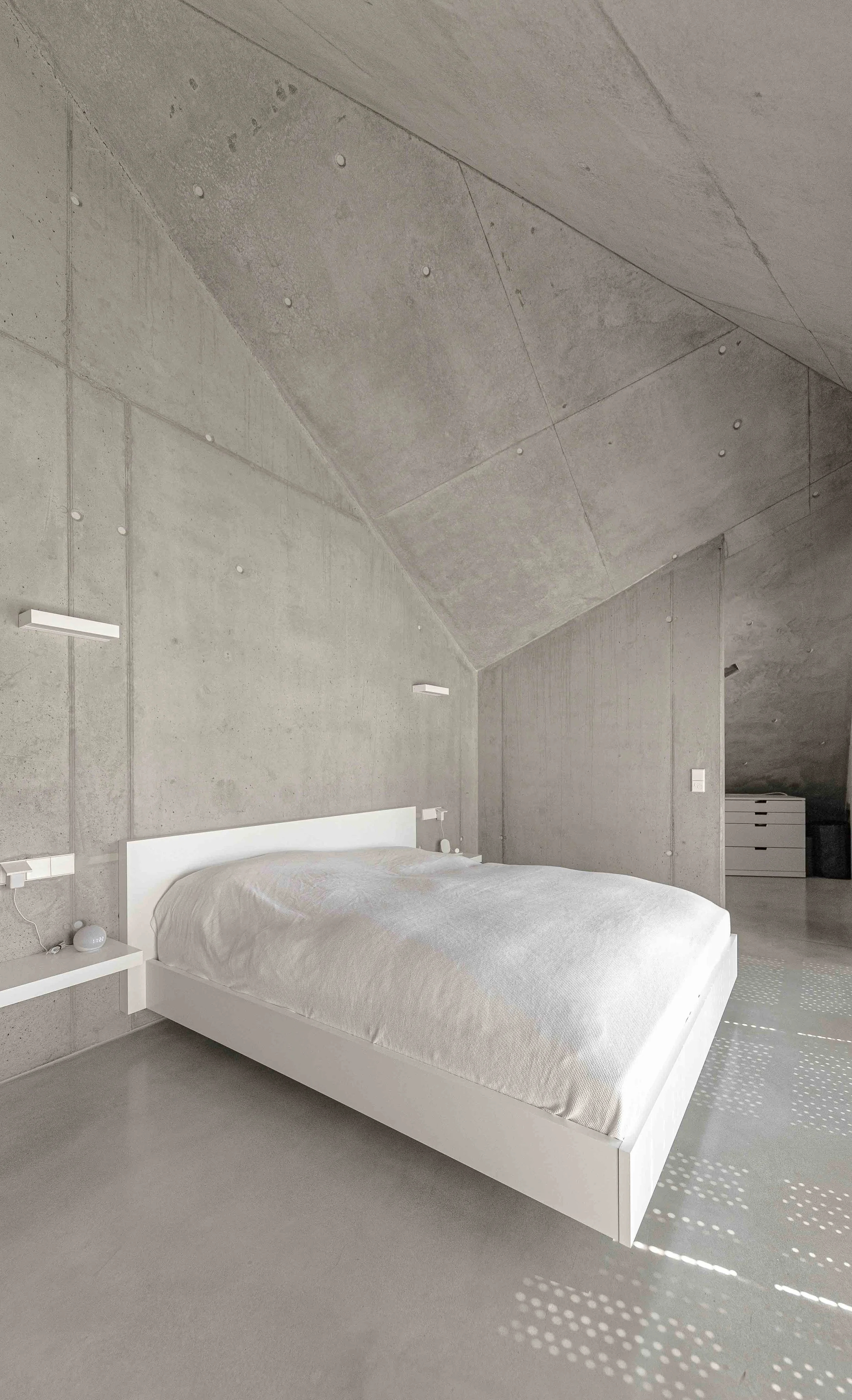 Schlafzimmer mit Betonwänden, weißem Bett und Nachttisch, Sonnenlicht fällt durch eine Fensteröffnung und schafft Musterschatten auf dem Boden.