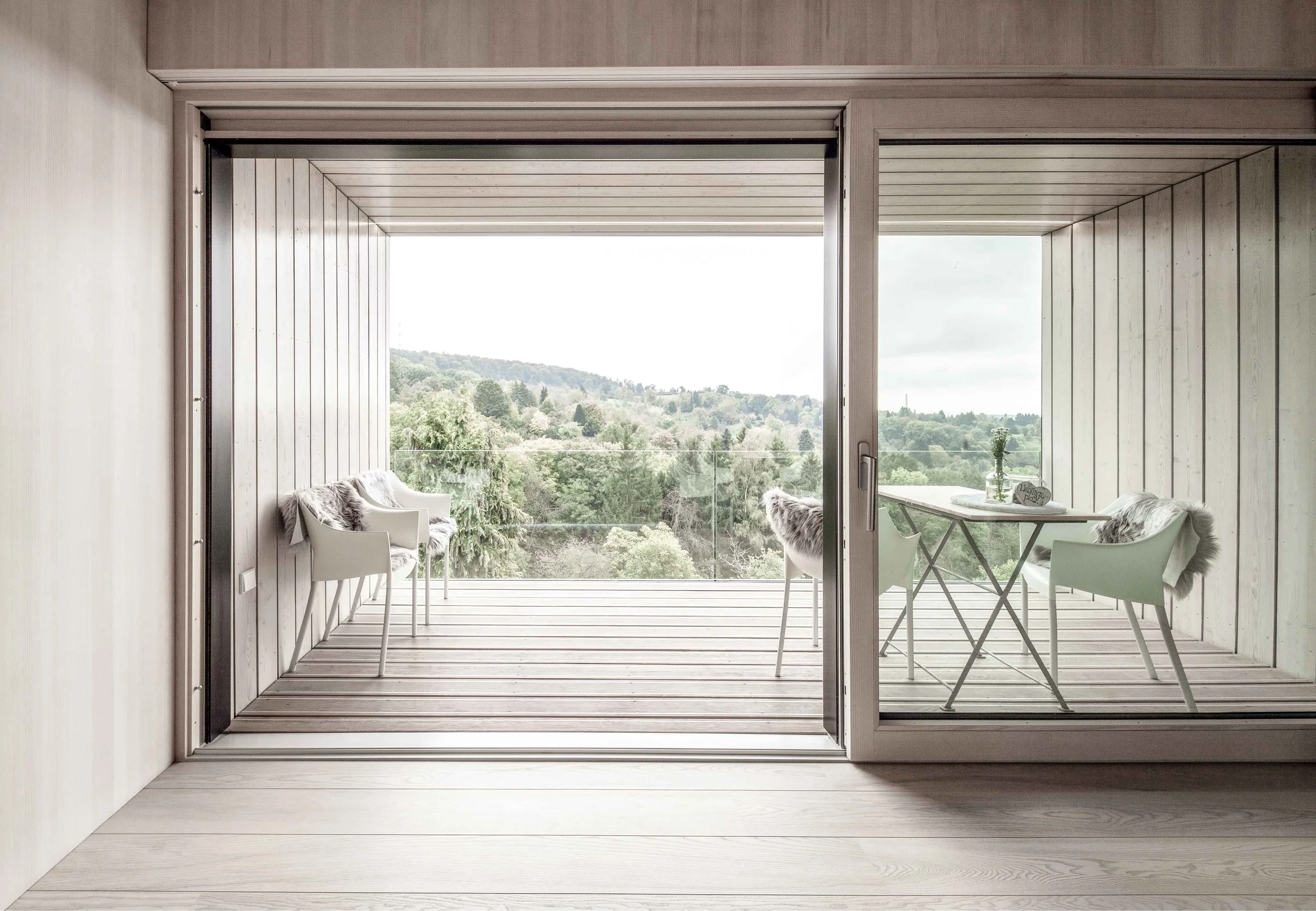haus GRO - Blick auf eine offene Terrasse mit weißen Stühlen, einem kleinen runden Tisch mit Dekoration, und einer großen Glasfront mit Blick auf eine grüne Landschaft mit Bäumen und Hügeln im Hintergrund.