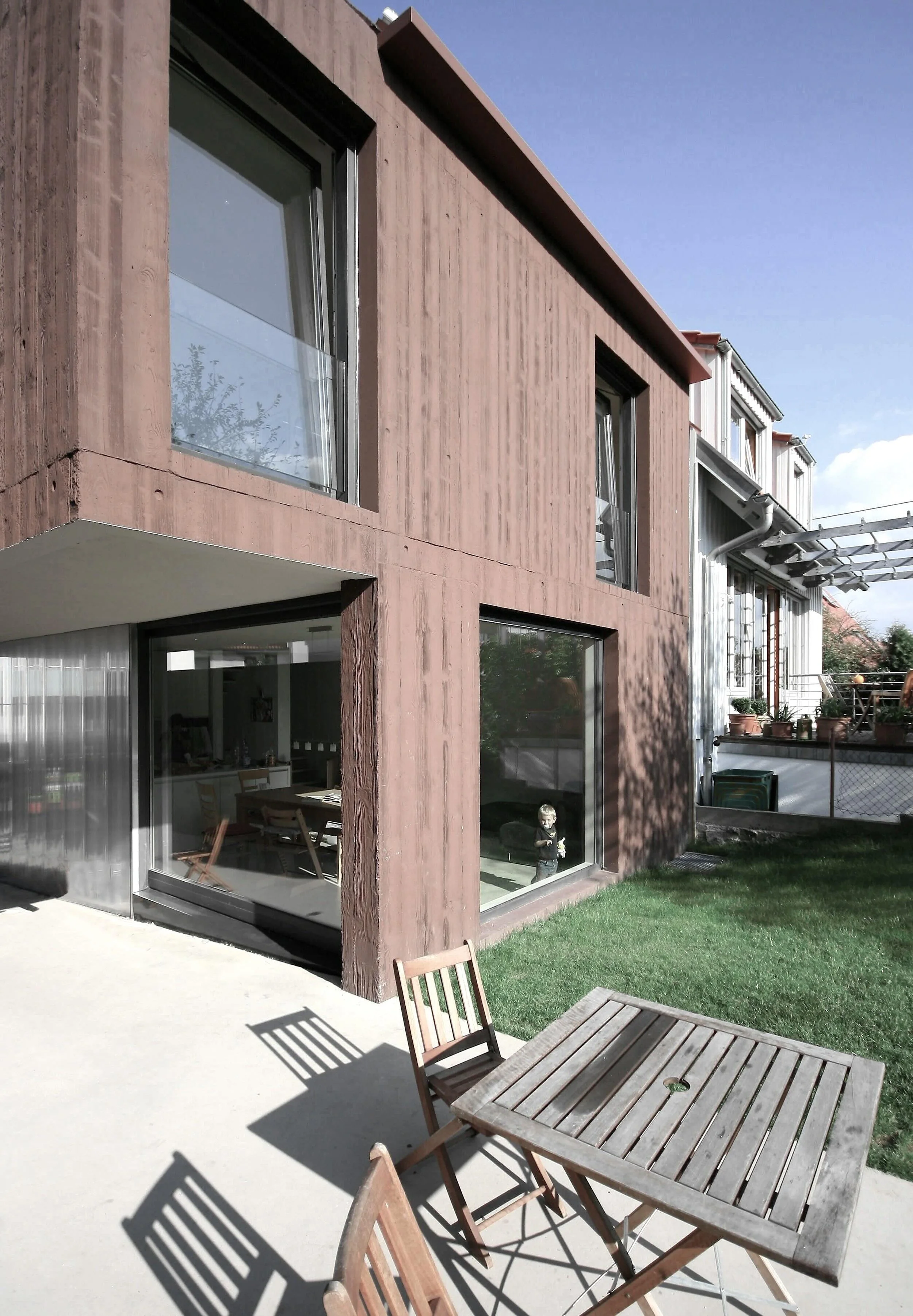 haus - B_Modernes Wohnhaus mit Sichtbetonfassade, großen Fenstern, Terrasse mit Holztisch und Stühlen sowie angrenzendem Garten bei Sonnenschein.