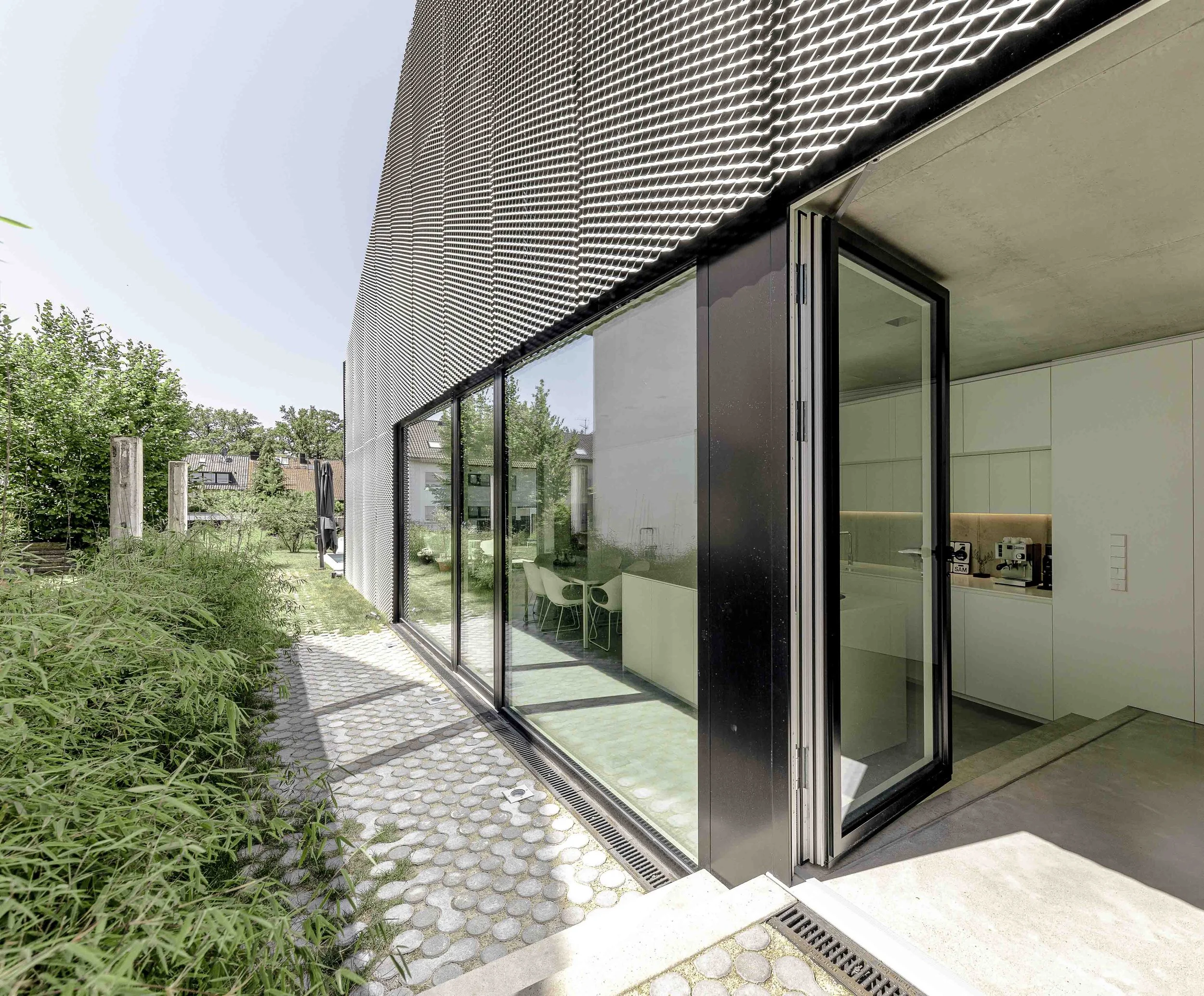 Moderne Gebäudefassade mit großen Glasfenstern und einer Metallfassade mit perforiertem Design, umgeben von grünen Büschen und einem gepflasterten Weg im Freien.