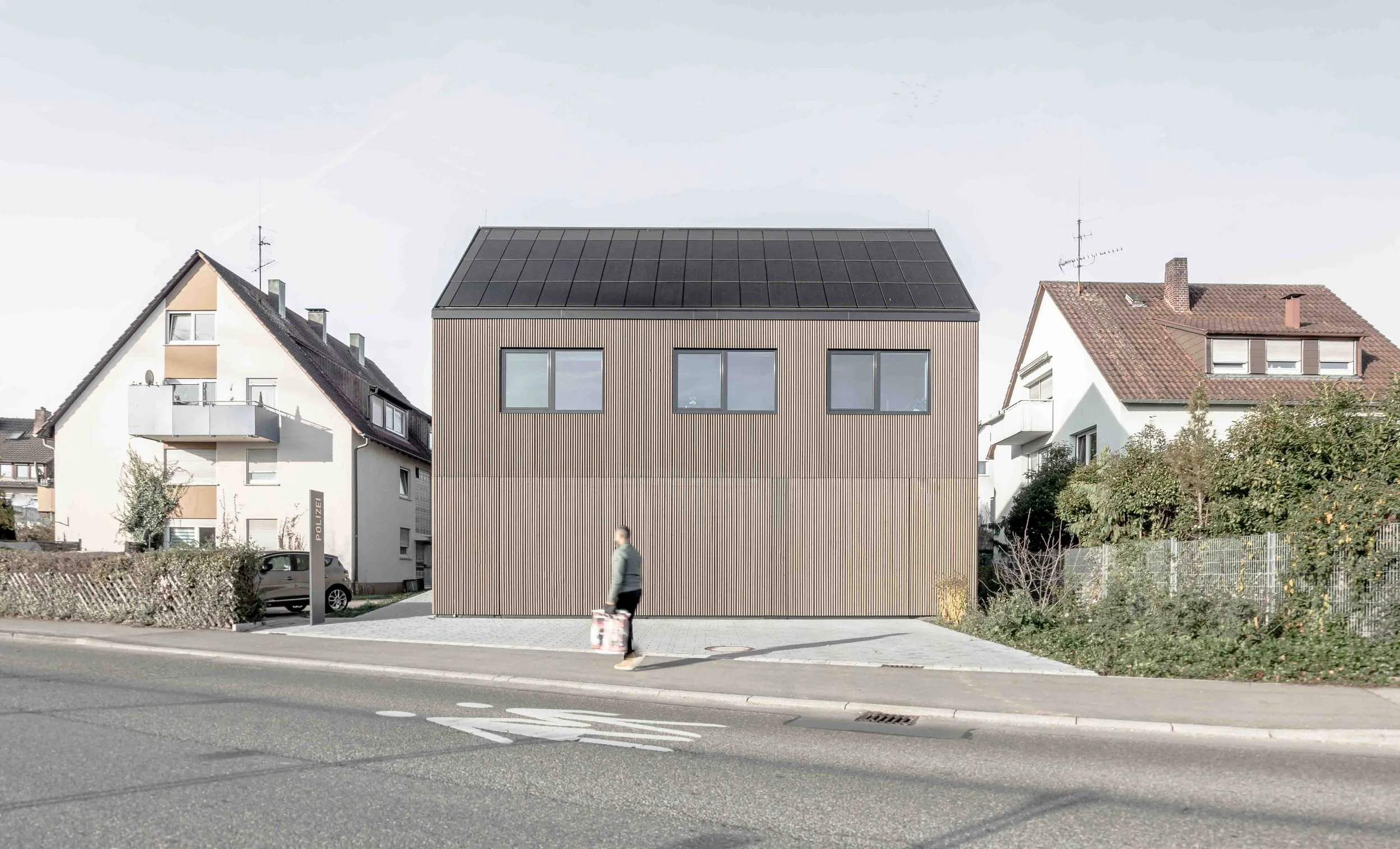 polizeiposten LE - Modernes, zweistöckiges Haus mit Holzfassade und großen Fenstern, umliegende alte Häuser mit roten Ziegeldächern, Straße mit Gehweg und parkendem Auto, eine Person läuft mit Handgepäck vorbei, blauer Himmel.