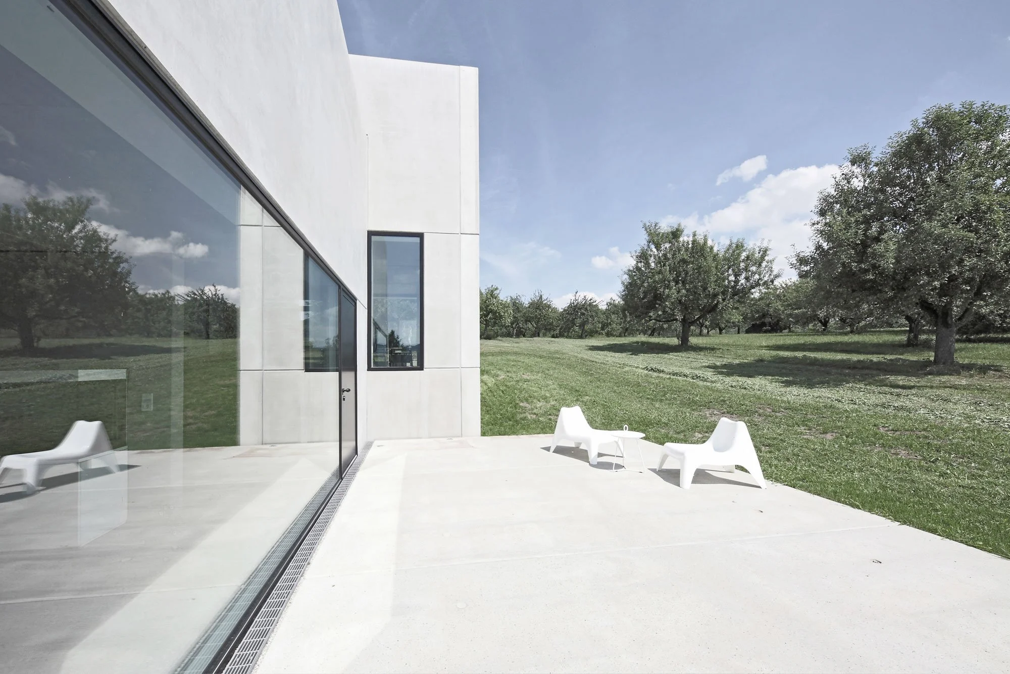 haus SG - Moderner whiteischer Bau mit großen Fensterflächen, Blick auf einen grünen Garten mit Bäumen, wenige weiße Gartenmöbel auf einer Terrasse.
