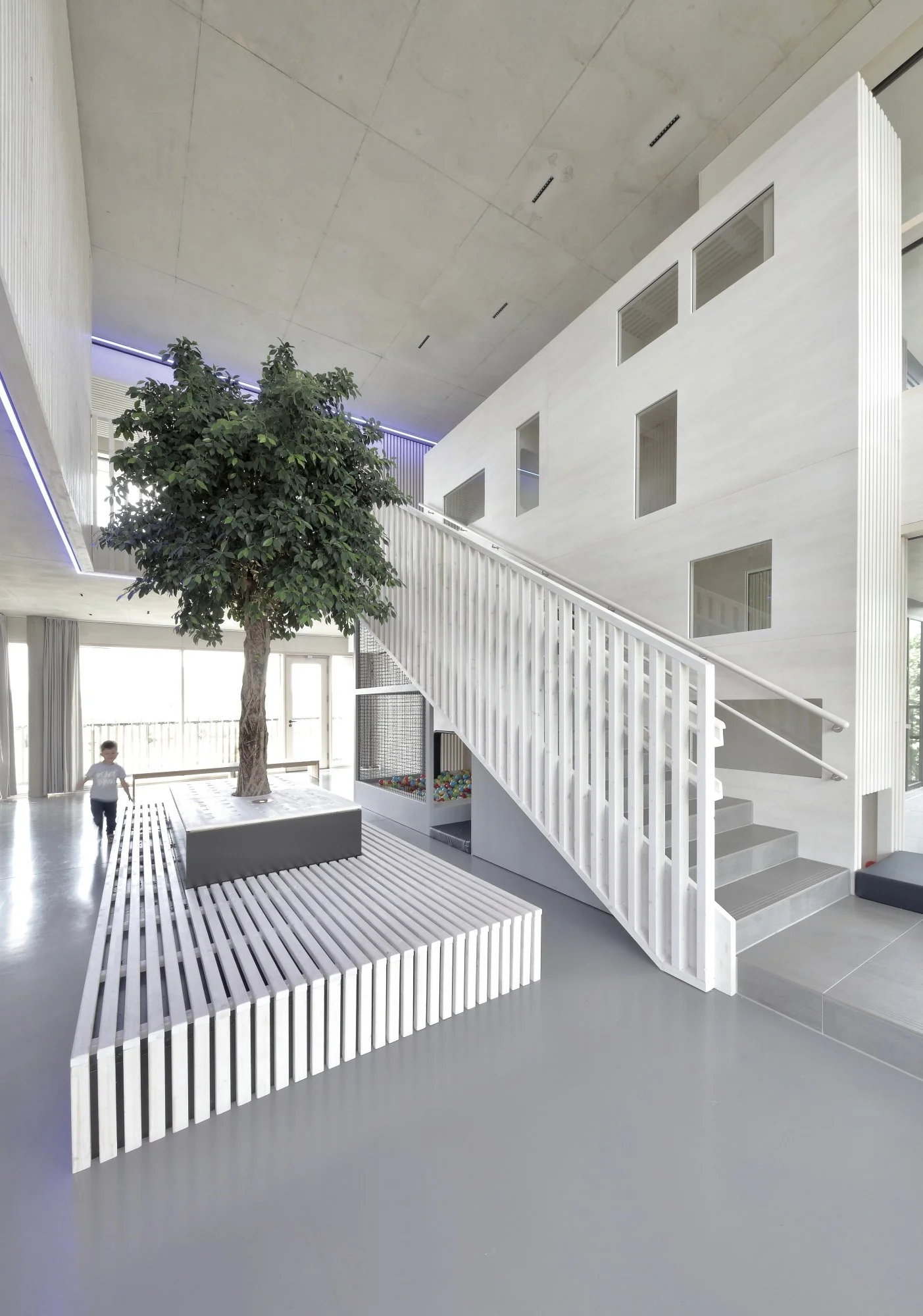 kinderhaus BUGA - Innenraum mit großem Baum in Topfpflanze, Treppe, weißen Holzdecke und modernen Fenstern. Im Vordergrund eine Sitzbank und ein Gitter mit bunten Bällen. Ein Kind läuft durch den Raum.
