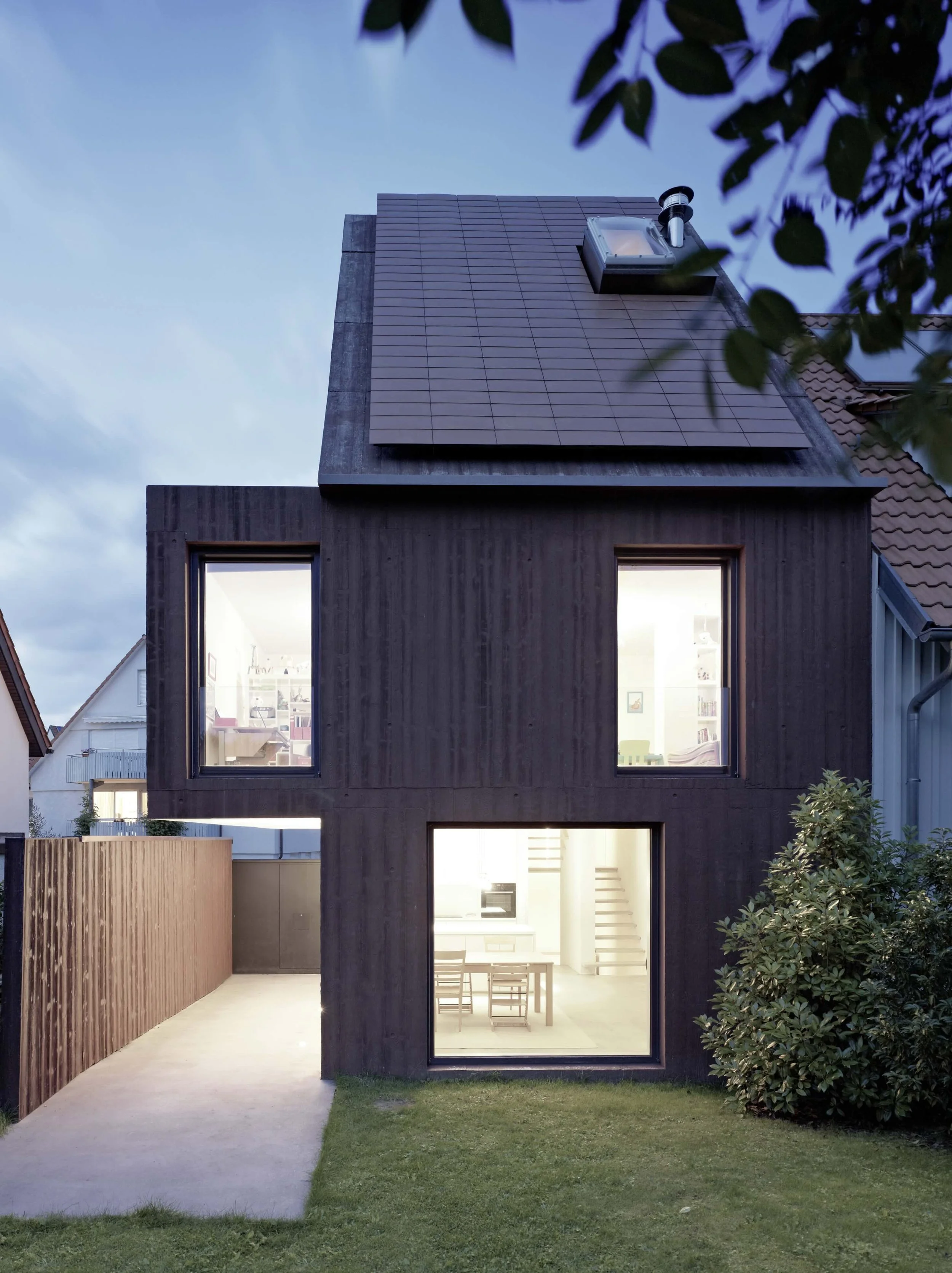 haus - B_Modernes schwarzes Einfamilienhaus mit großen Fenstern, begrüntem Vorgarten und sichtbarem Kamin unter blauem Abendhimmel.