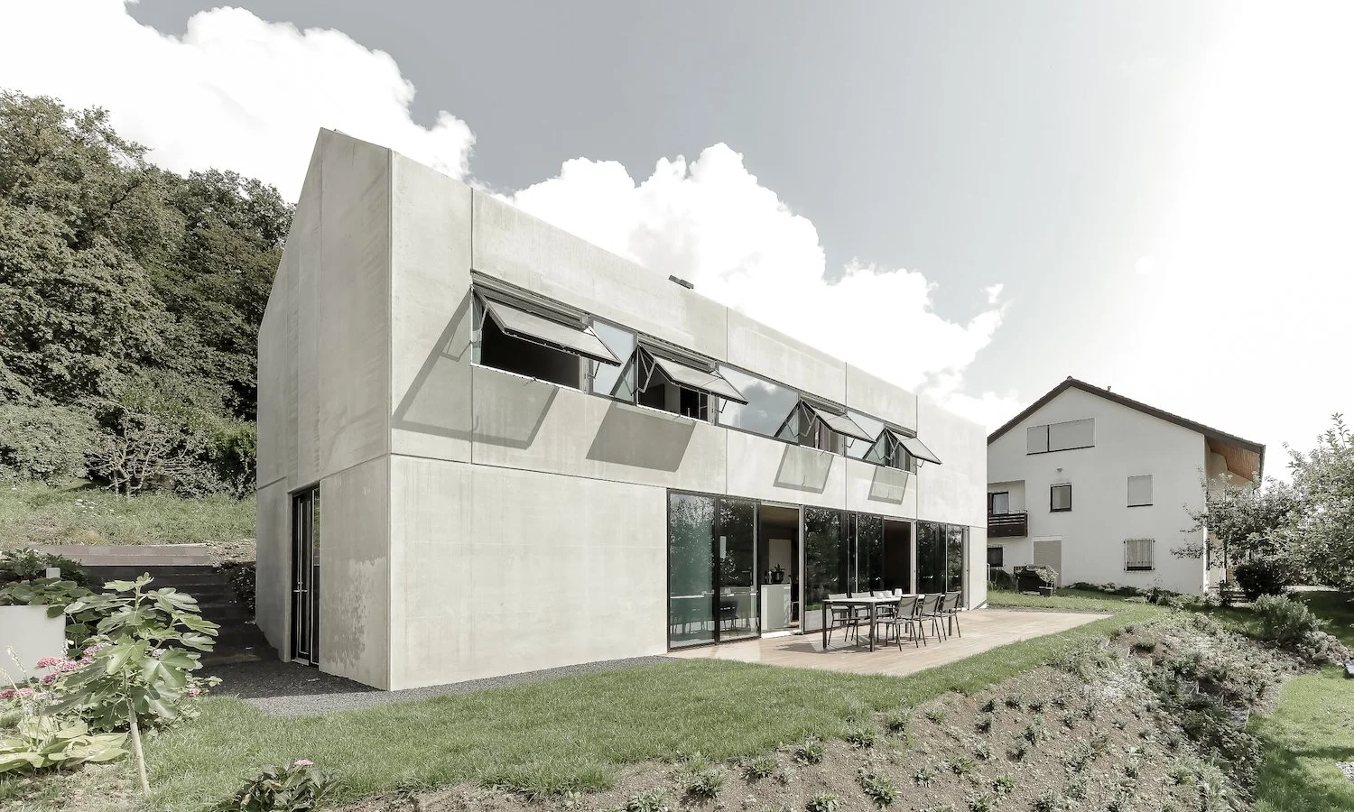 haus WÄ - Modernes zweistöckiges Haus mit großen Glasfenstern und Sonnenschutzmarkisen auf der Terrasse, umgeben von Garten und Natur.