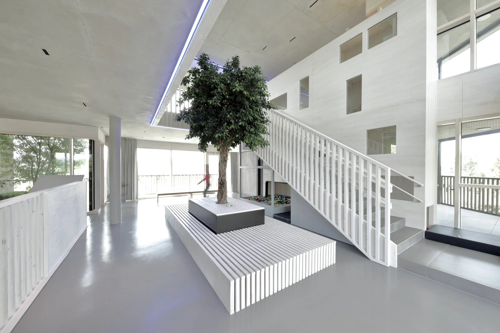 kinderhaus BUGA - Innenansicht eines modernen, hell erleuchteten Gebäudes mit weißem und grauem Design. Im Mittelpunkt steht ein großer Baum in einem rechteckigen Pflanzkübel. 