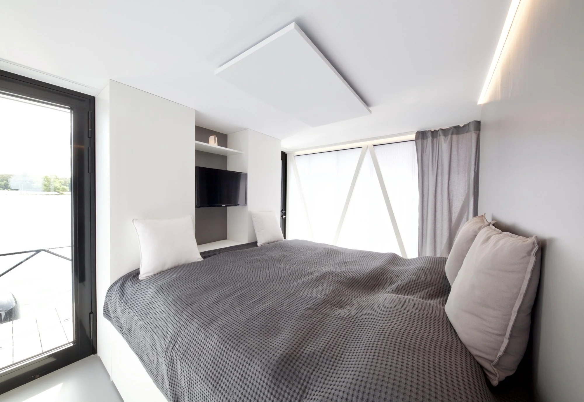 Modernes Schlafzimmer mit großem Bett, weißen Kissen und Bettdecke, großen Fenstern mit Vorhängen, Fernseher an der Wand, und geschmackvoller Deckenbeleuchtung.