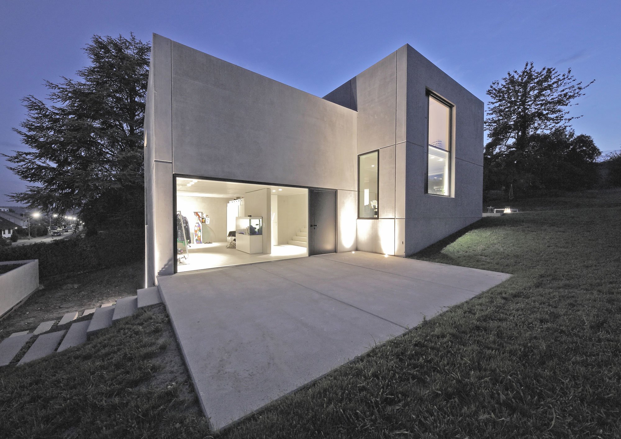 haus SG - Modernes, zweistöckiges Haus bei Nacht mit offener Garage, großen Fenstern und außen beleuchteter Fassade, umgeben von Bäumen und Gras.