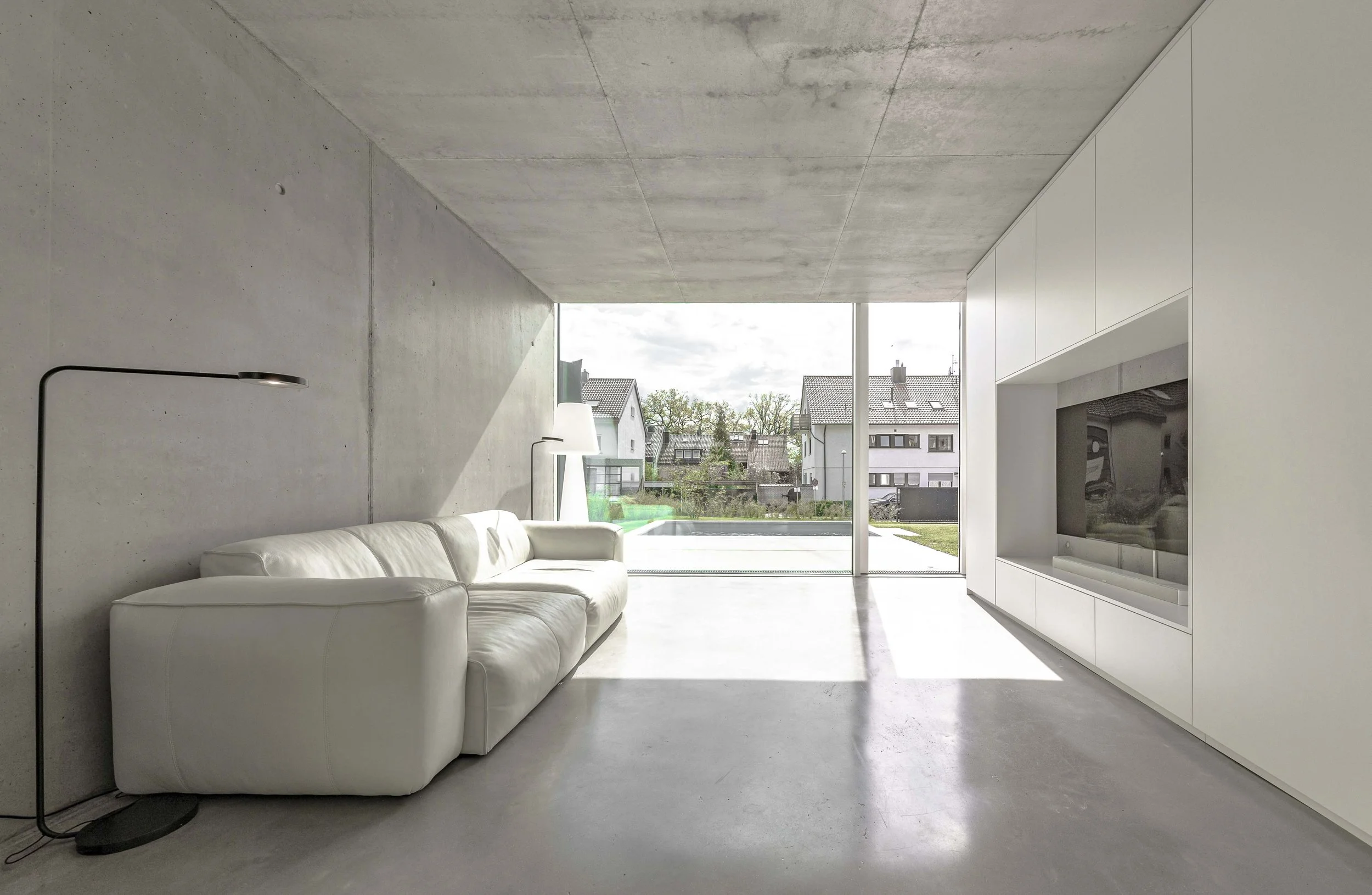 Moderner Wohnraum mit einer weißen Ledercouch, einem schwarzen Stehlampe, einem großen Fernseher in einem weißen Einbauschrank und einer großen Fensterfront mit Blick auf Nachbarhäuser und Bäume.
