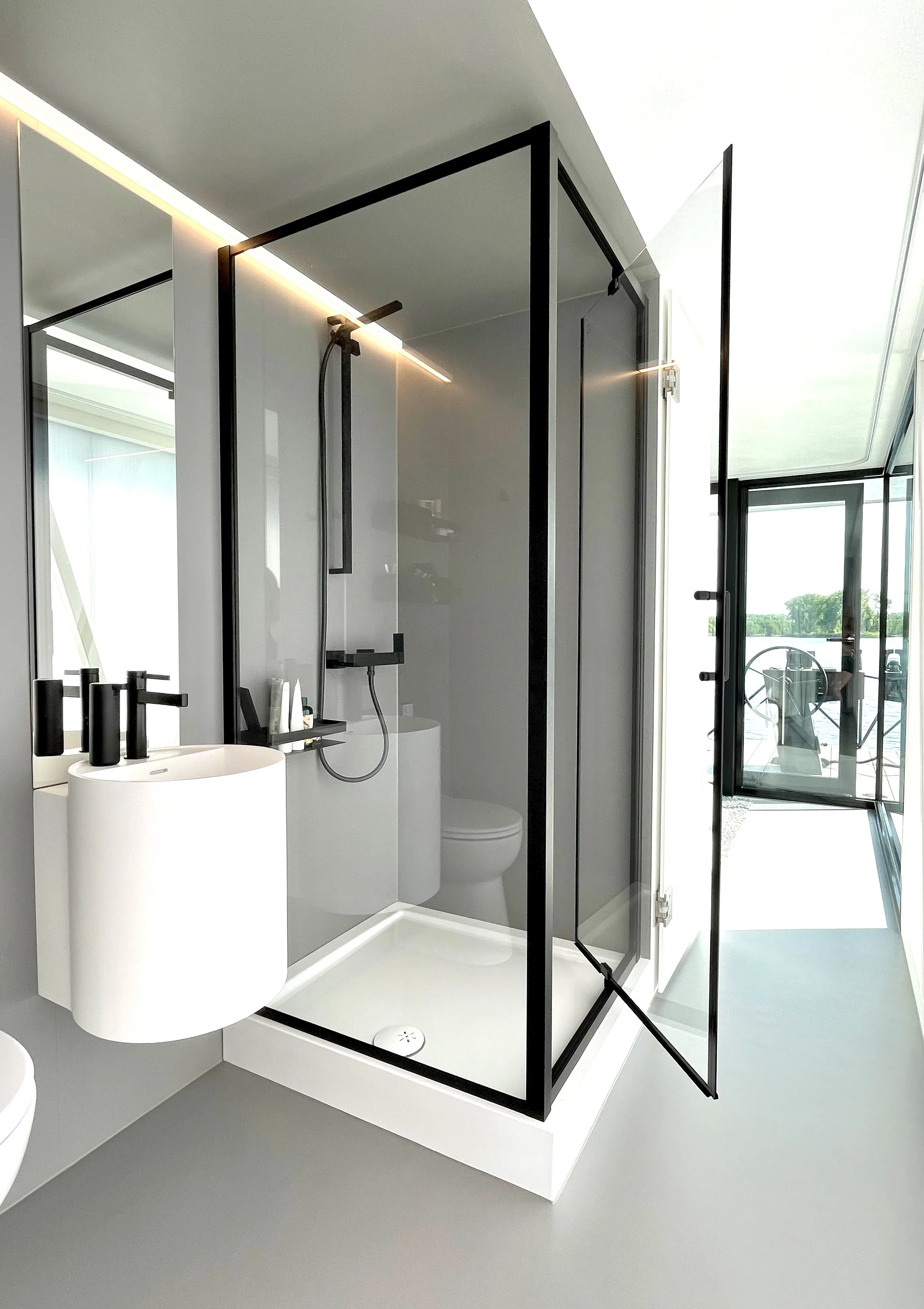 Modernes Badezimmer mit Dusche, Wandspiegel, Waschbecken und Toilette, Blick auf eine Terrasse mit Stühlen im Hintergrund.